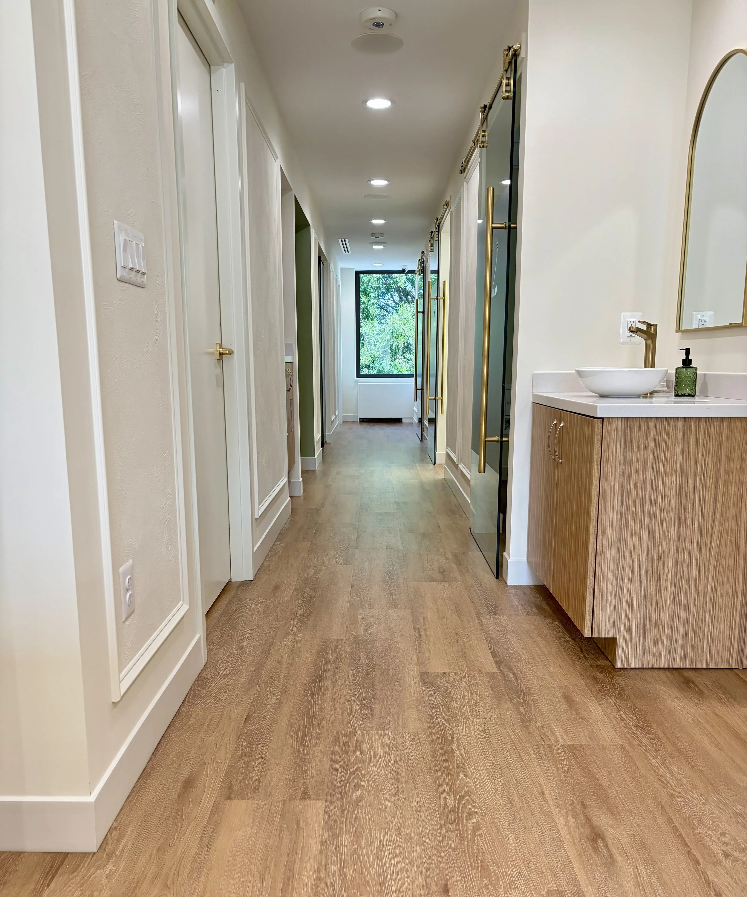Modern hallway with wooden floors, white walls, and gold accents, leading to a room with a large window showing green trees outside. On the right, there is a small sink with a white vessel basin, gold faucet, and mirror with gold frame above.