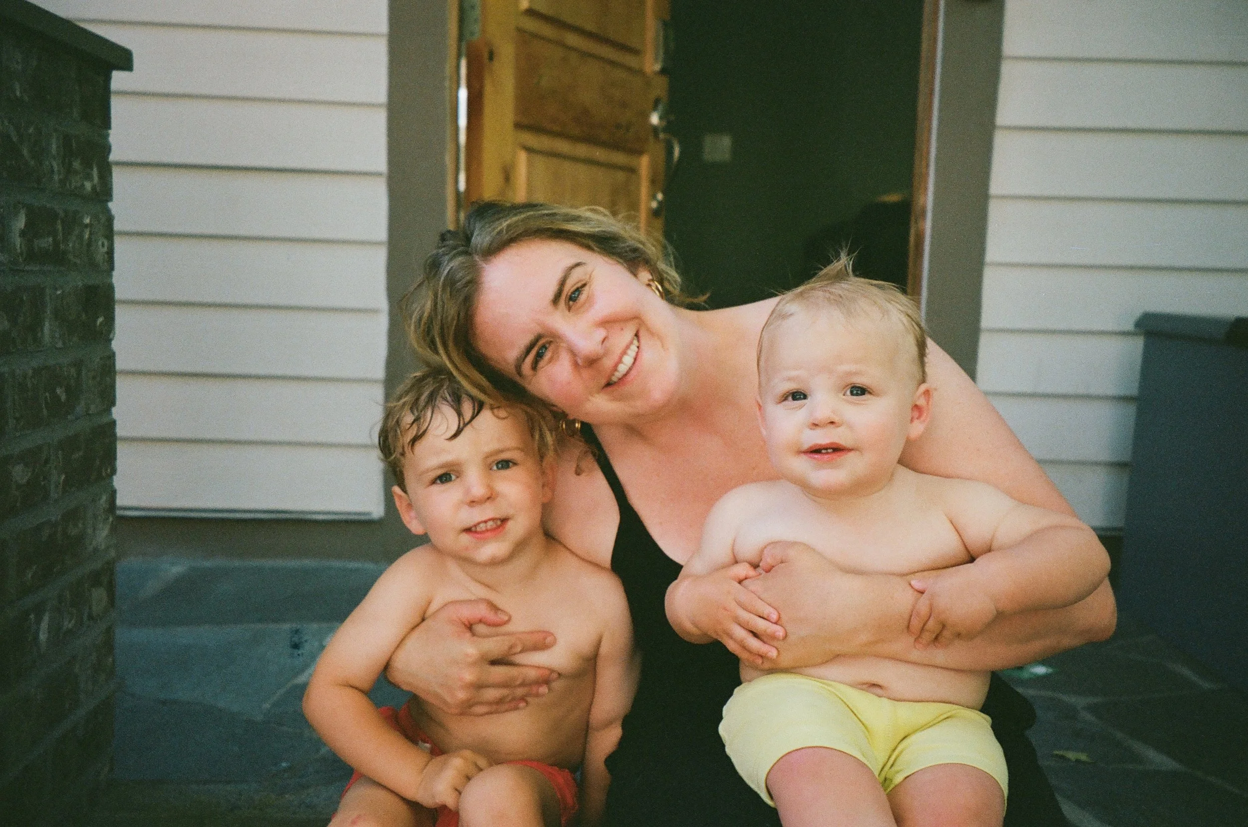 Happy woman with short hair, smiling, holding two young boys, all shirtless, outdoors on a porch near a brick wall and a garage door.