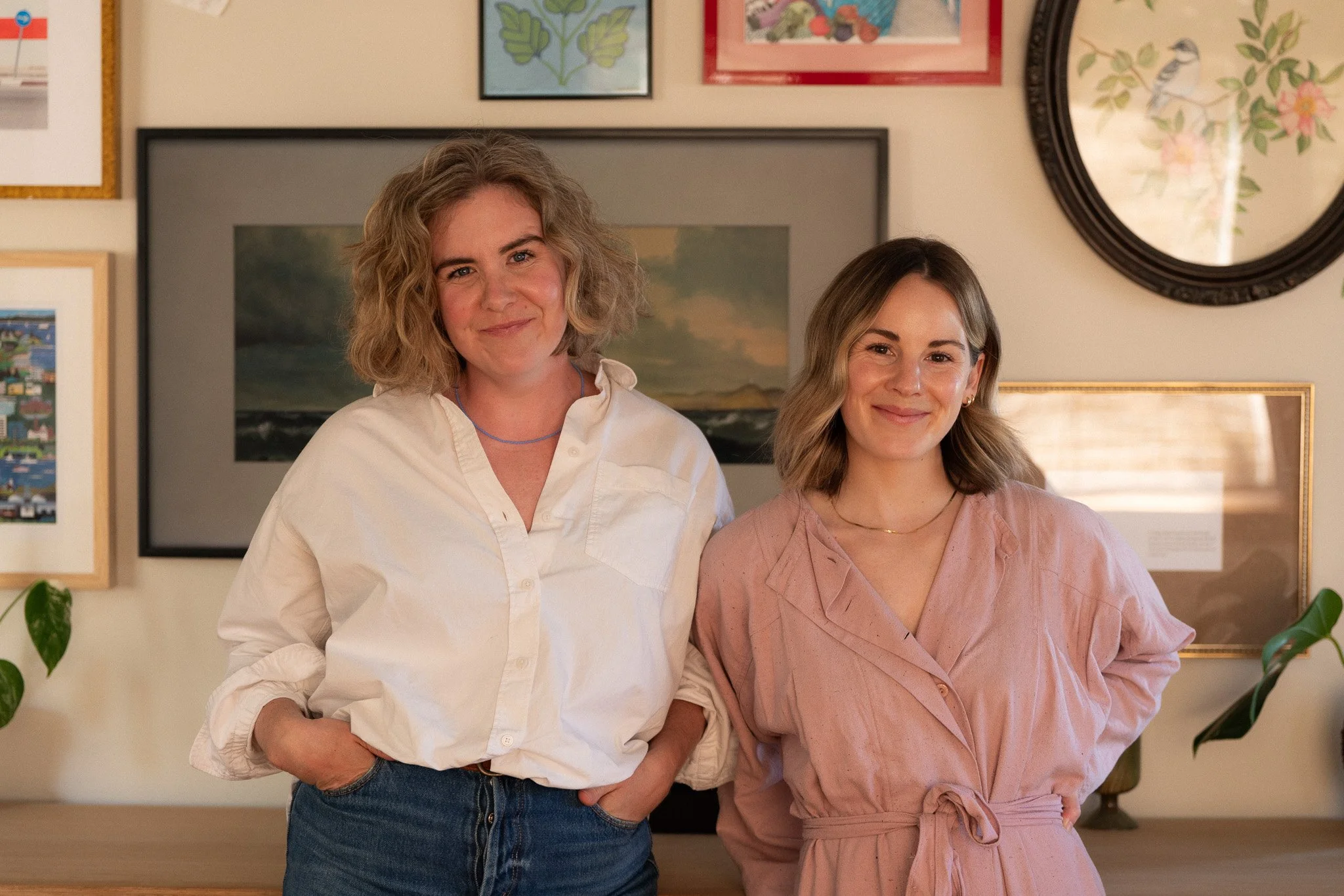 Two women standing together in a room with picture frames on the wall behind them. The woman on the left has curly blonde hair, wearing a white shirt, and has her hand in her pocket. The woman on the right has straight brown hair, wearing a light pink dress, with her arm around the other woman.