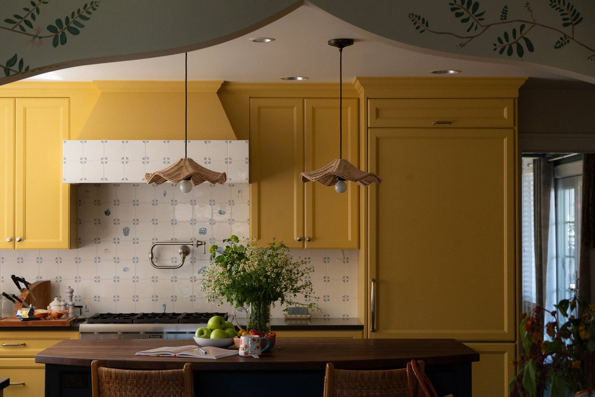 Kitchen with yellow cabinets, a white tiled backsplash, two hanging pendant lights with fabric shades, a bowl of green apples, an open book, and floral decorations visible on the ceiling and side.