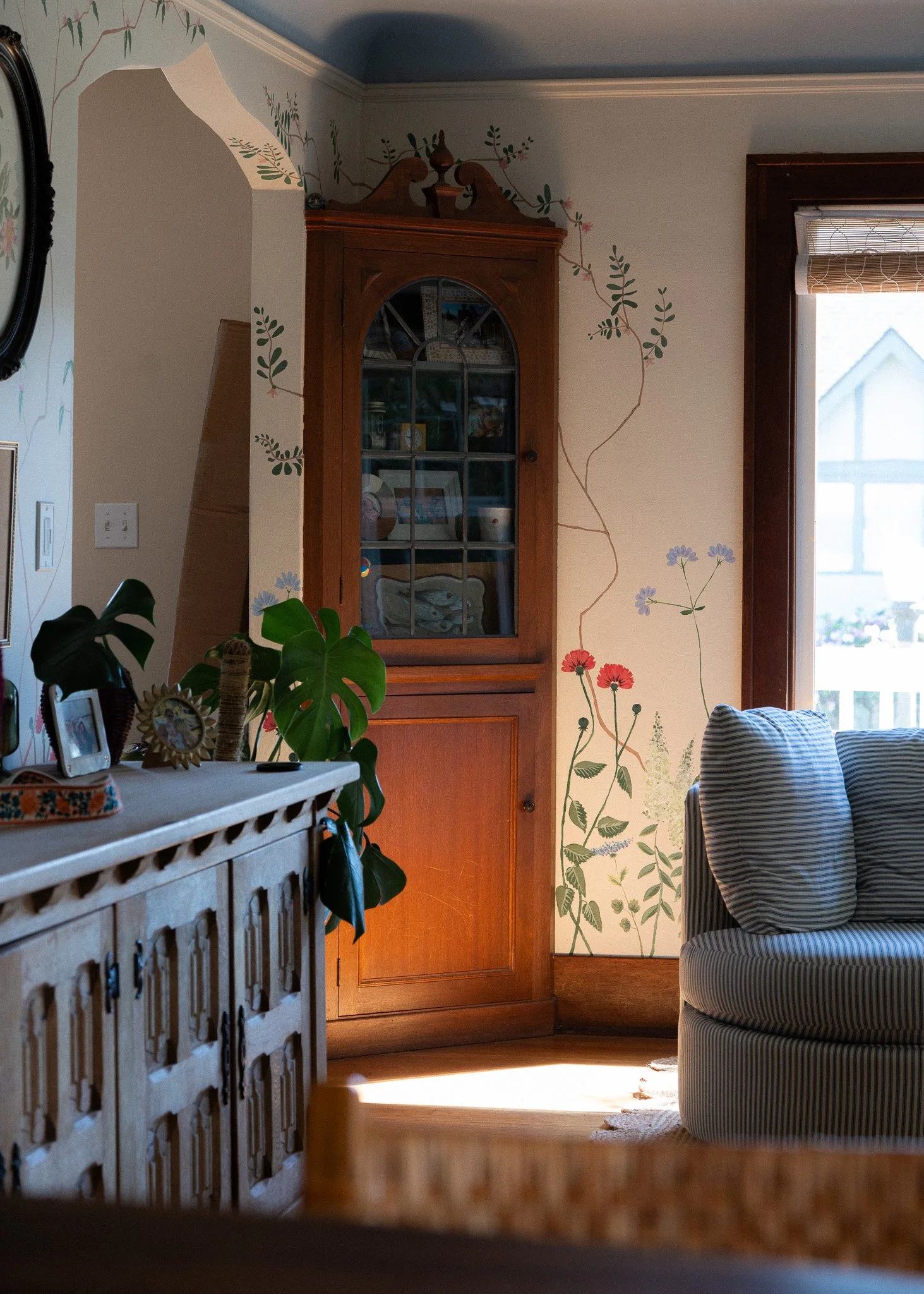 A corner of a living room featuring a wooden cabinet with glass doors, a striped armchair, a painted floral mural on the wall, and a window with blinds letting in natural light.