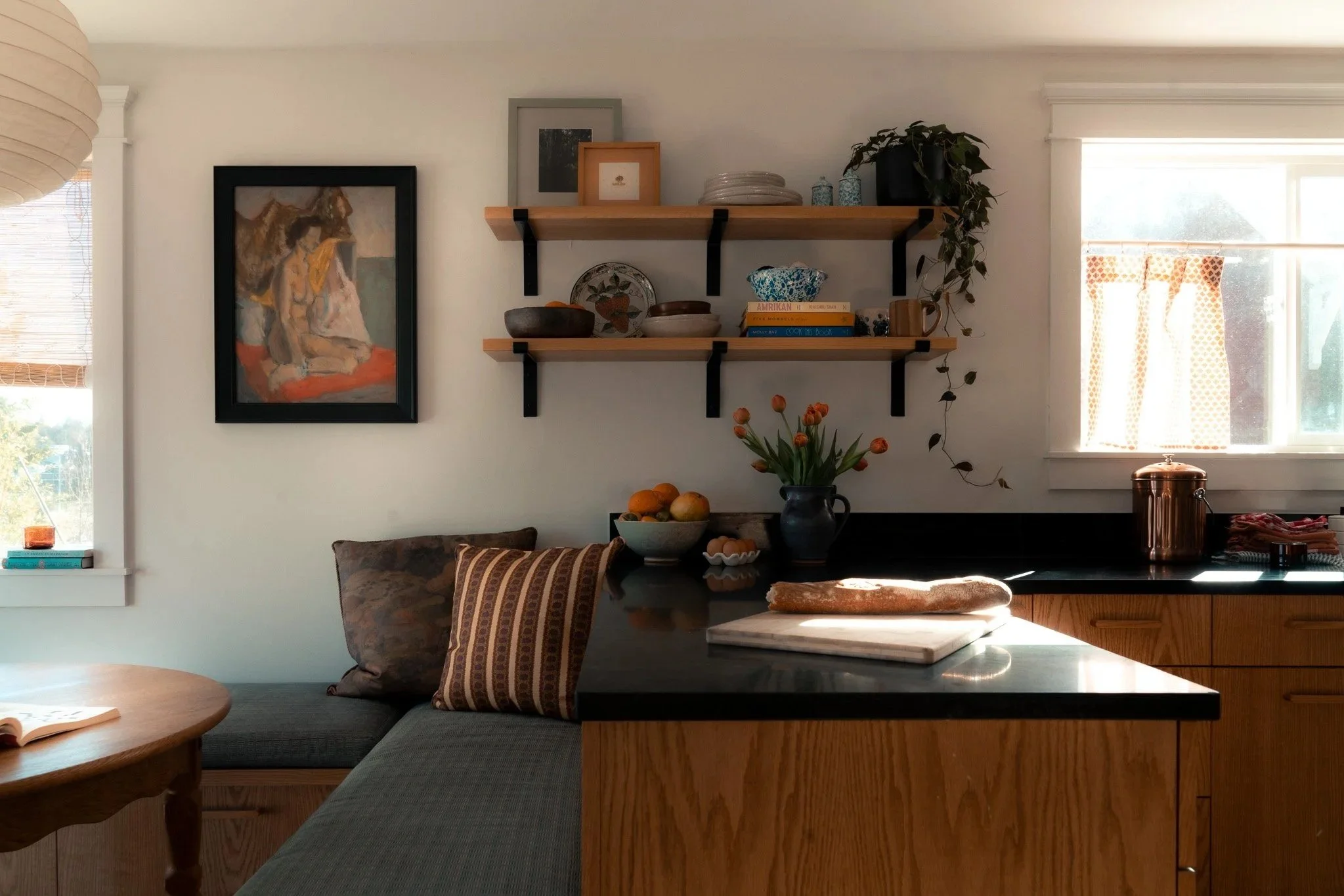 A cozy kitchen and living space with wooden shelves holding bowls and decorative items, a framed painting of a lion, a window with blinds and sunlight, a couch with a striped cushion, a bowl of oranges, and a marble countertop with a loaf of bread.