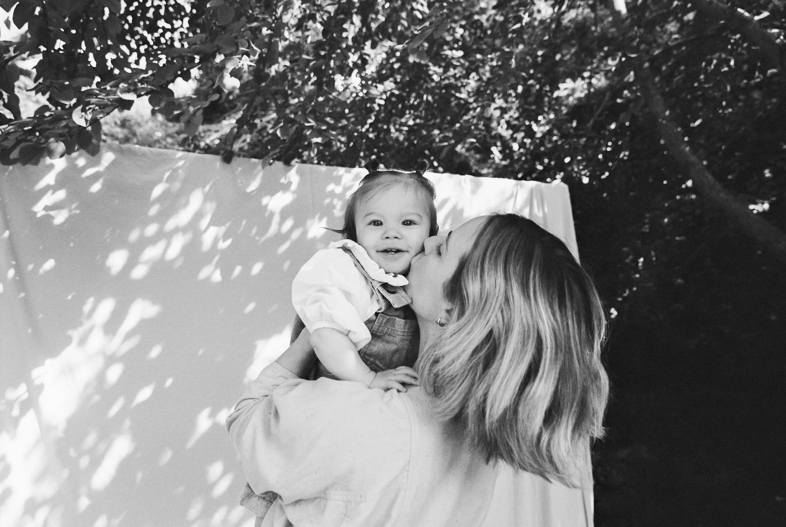 A woman with long blonde hair holding a young girl with pigtails, outdoors under a tree with light filtering through the leaves.