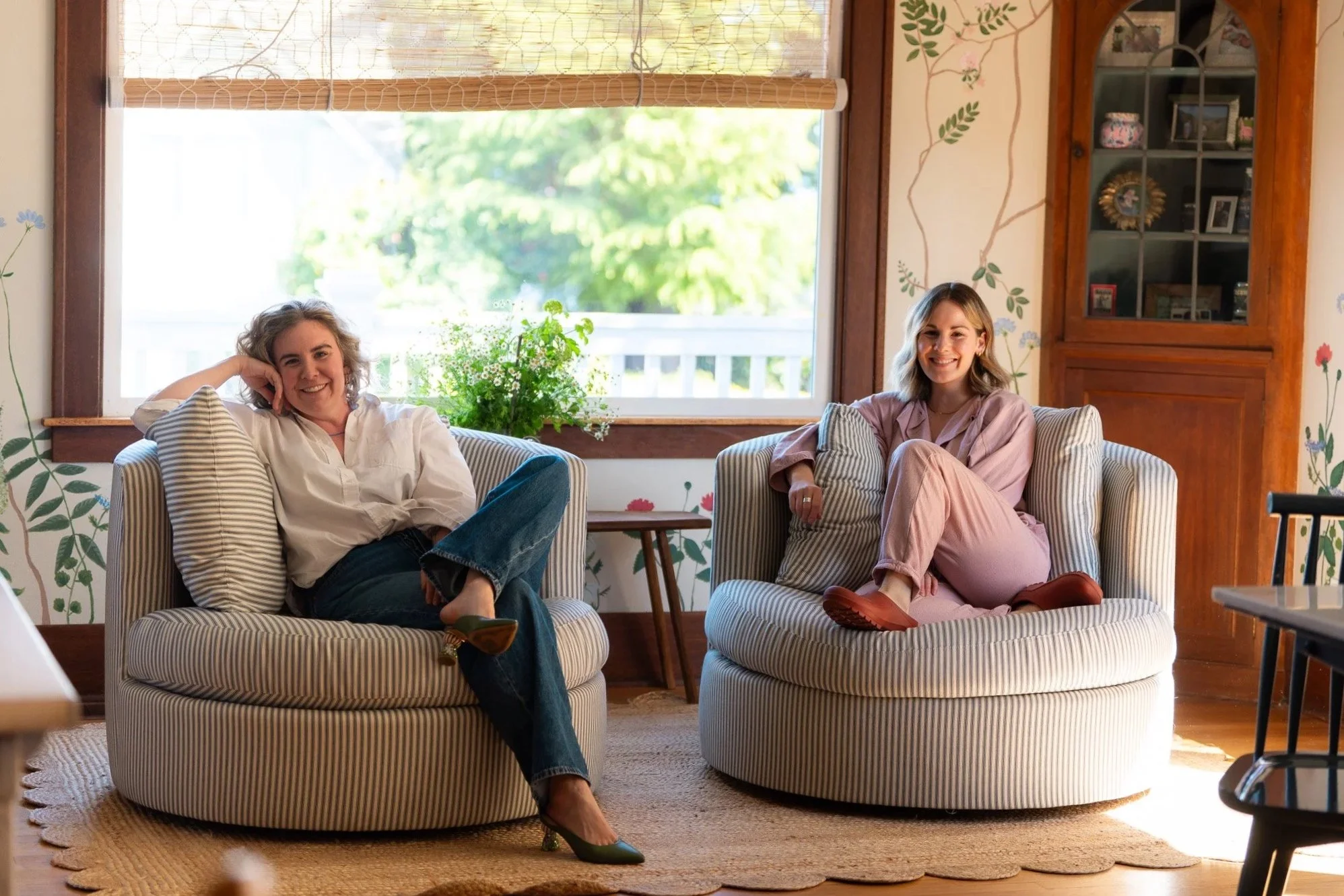 Two women sitting on striped armchairs in a cozy, sunlit room with floral wall art and wooden furniture, smiling and relaxing.