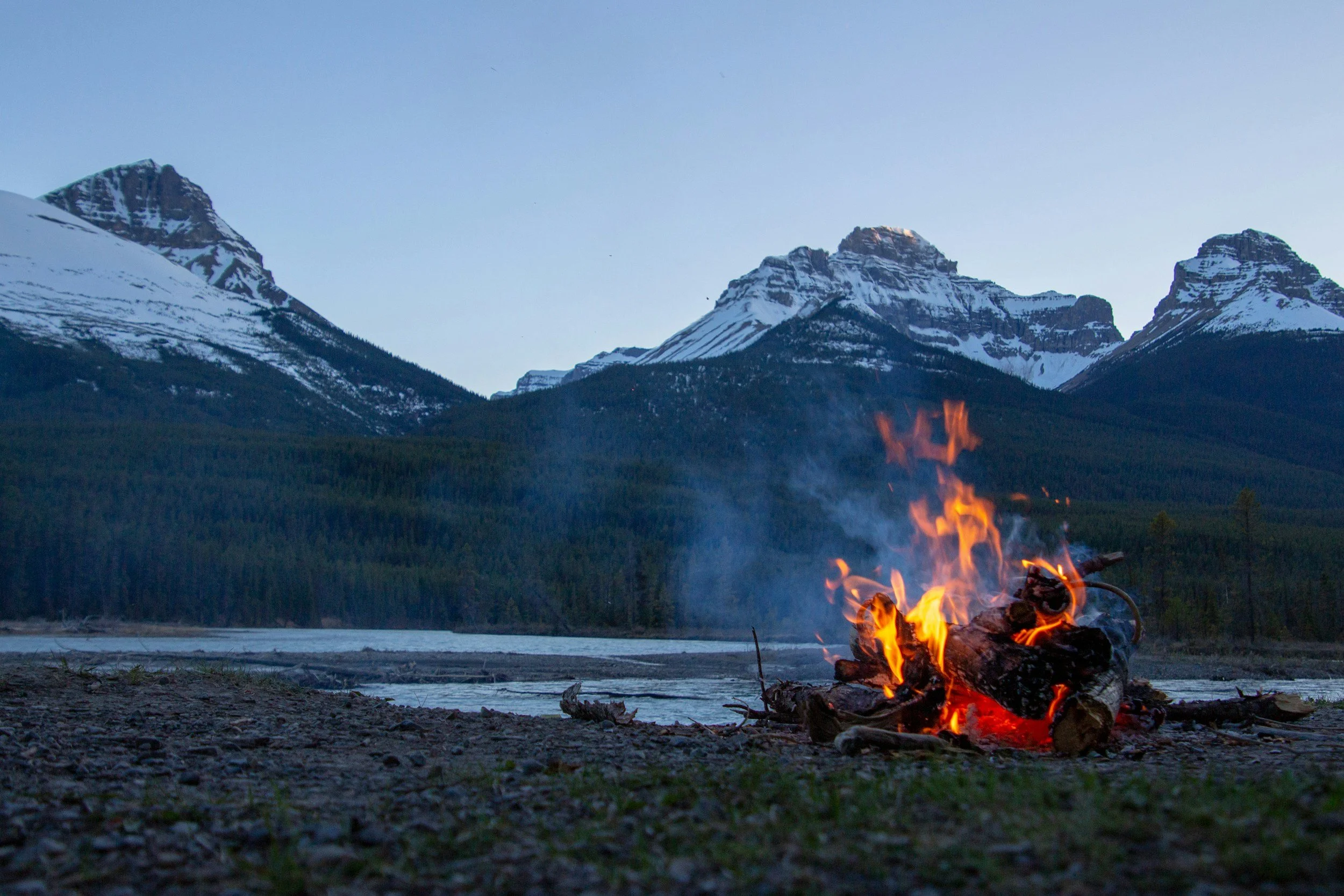 Campfire burning on rocky shore with snow-capped mountains in the background and a clear blue sky.