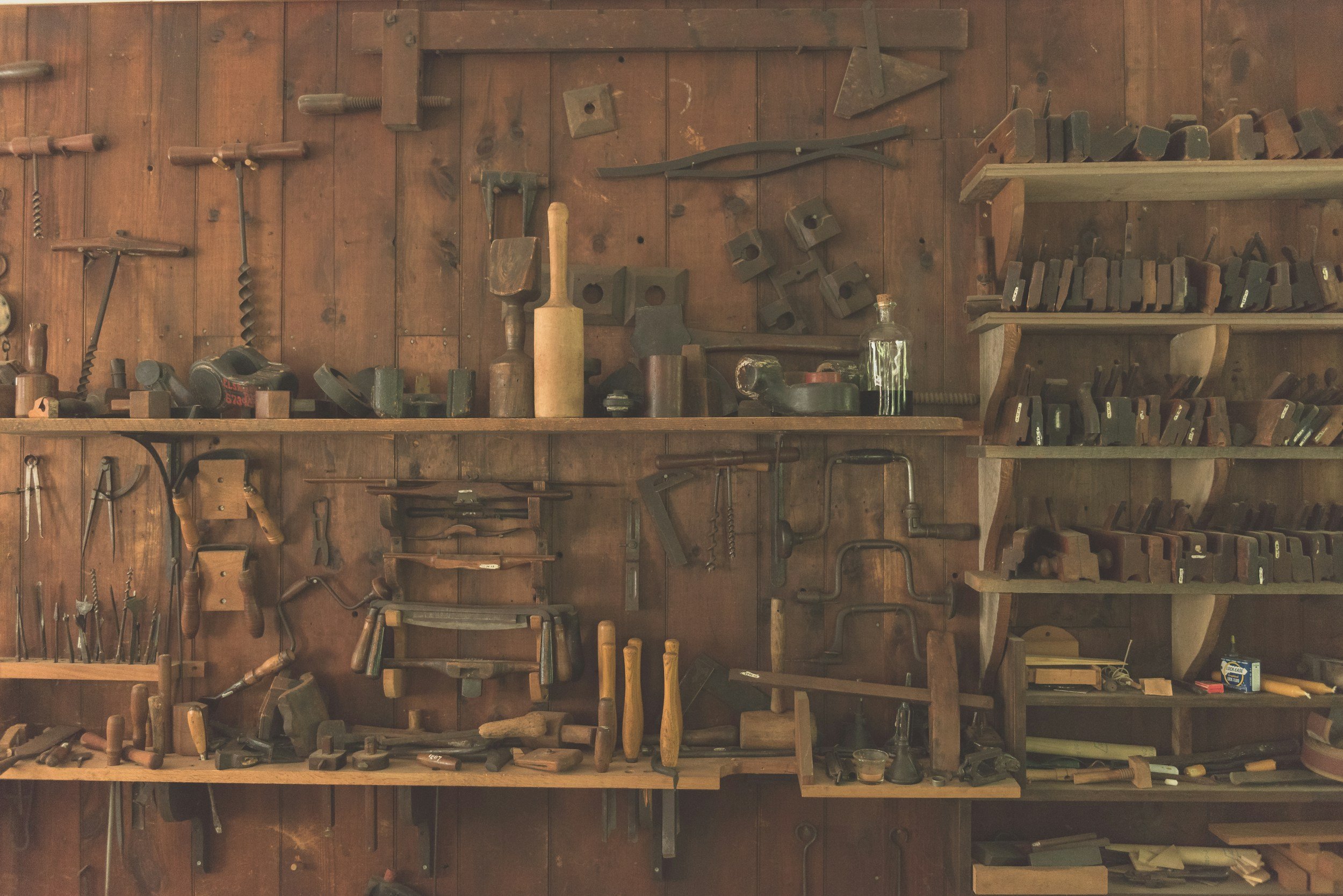 A wooden wall with various antique hand tools such as hammers, chisels, and woodworking tools arranged and stored on shelves and hooks.