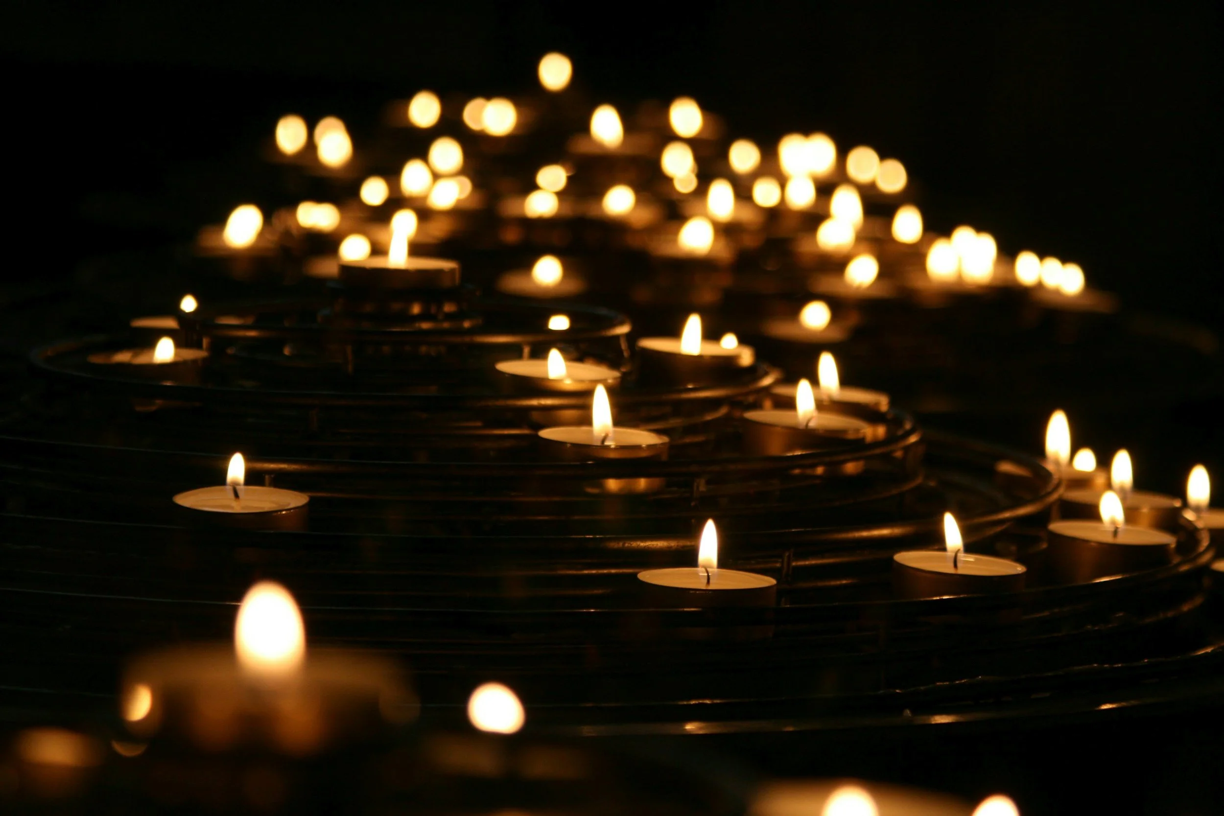 Multiple small tea light candles burning in a dark setting, arranged in a pattern with some candles out of focus in the background.