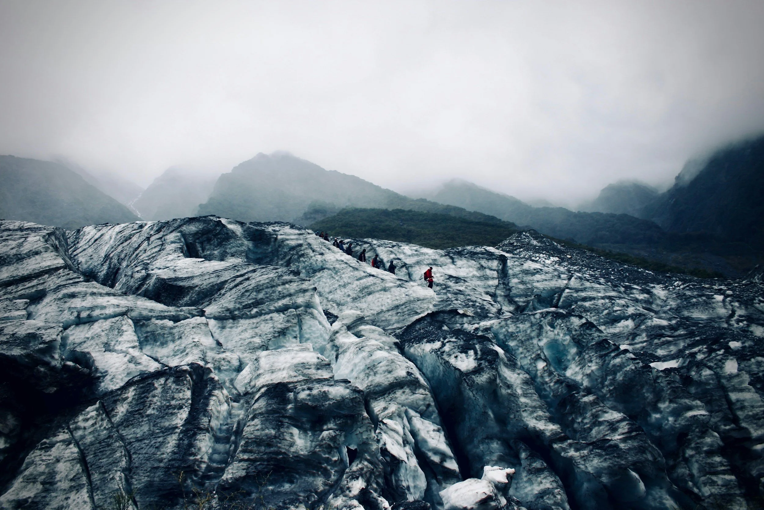 A glacier with a group of hikers walking on it, surrounded by mountains and foggy weather.