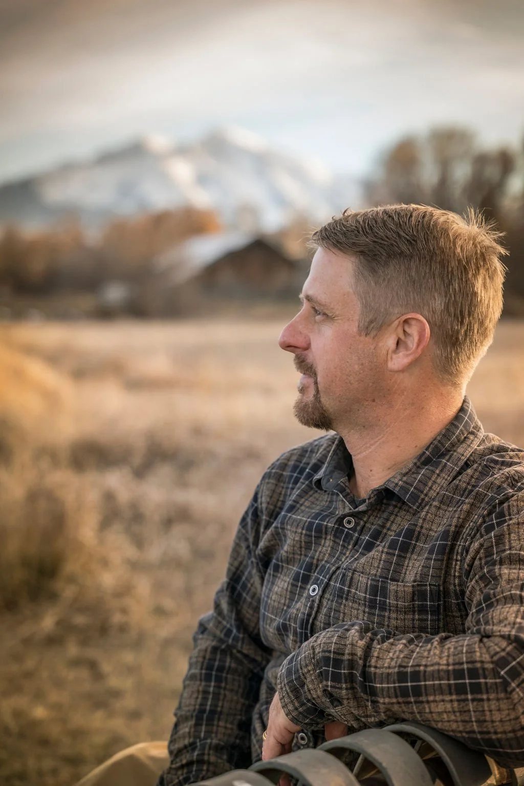 A man sitting outdoors in a rural landscape with snow-capped mountains in the background, gazing to the side.