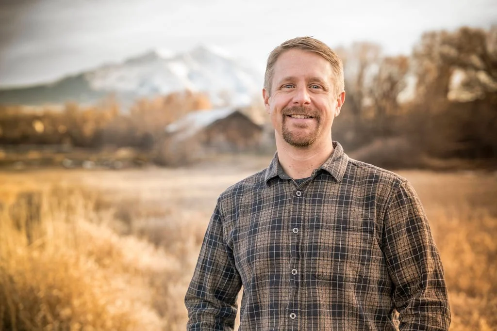 A smiling man with short blonde hair and a beard, wearing a plaid shirt, standing outdoors near a marshland, with snow-capped mountains in the background.