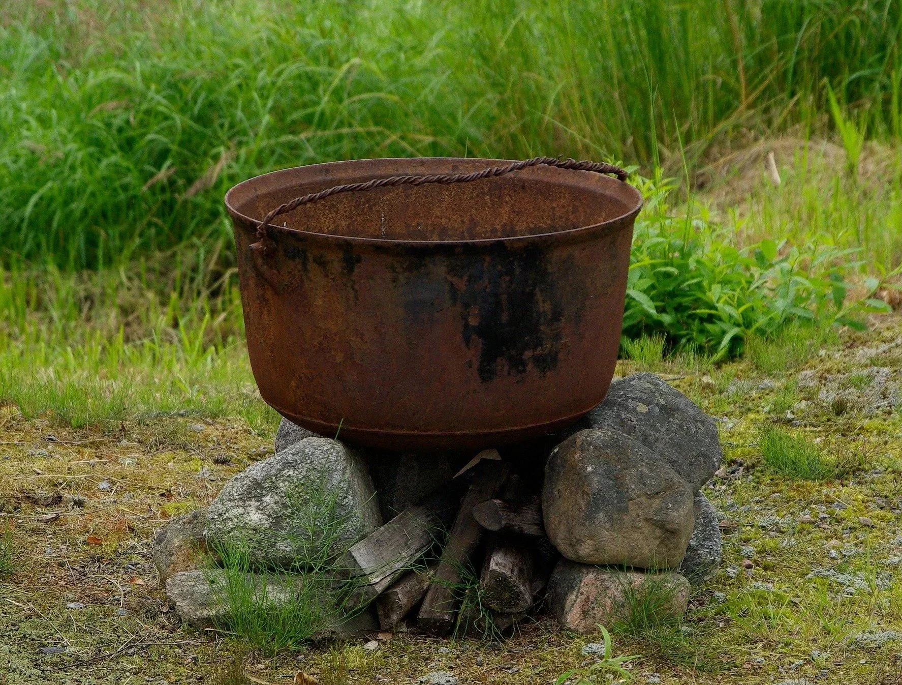 An old, rusty metal bucket resting on a stack of rocks outdoors amid green grass and plants.
