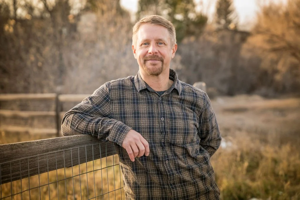 A man with short blond hair, a beard, and a mustache, wearing a plaid shirt, leaning against a wooden fence outdoors during sunset with trees in the background.