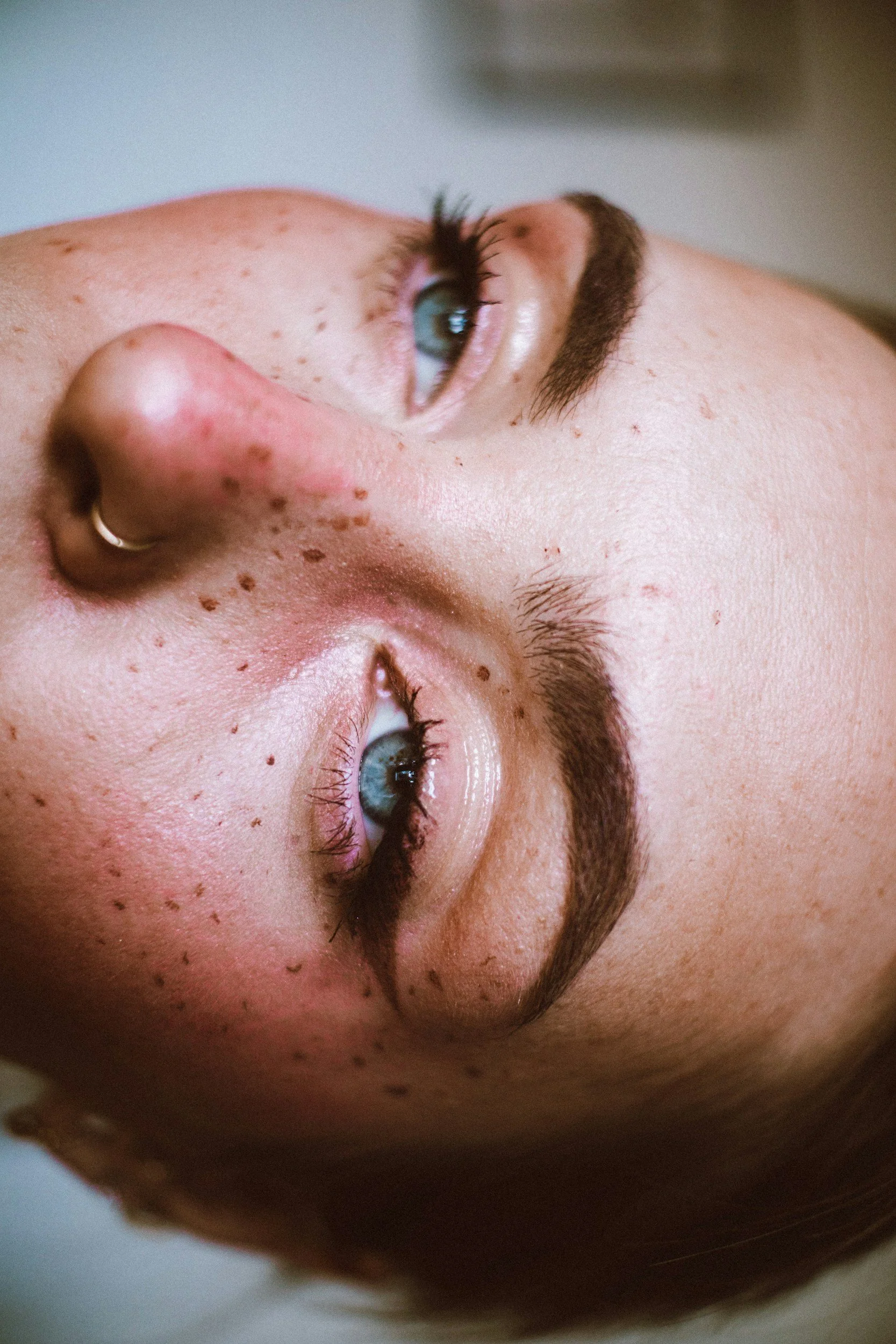 Close-up of a person's face with blue eyes, dark eyebrows, freckles, and a nose piercing, seen upside down.