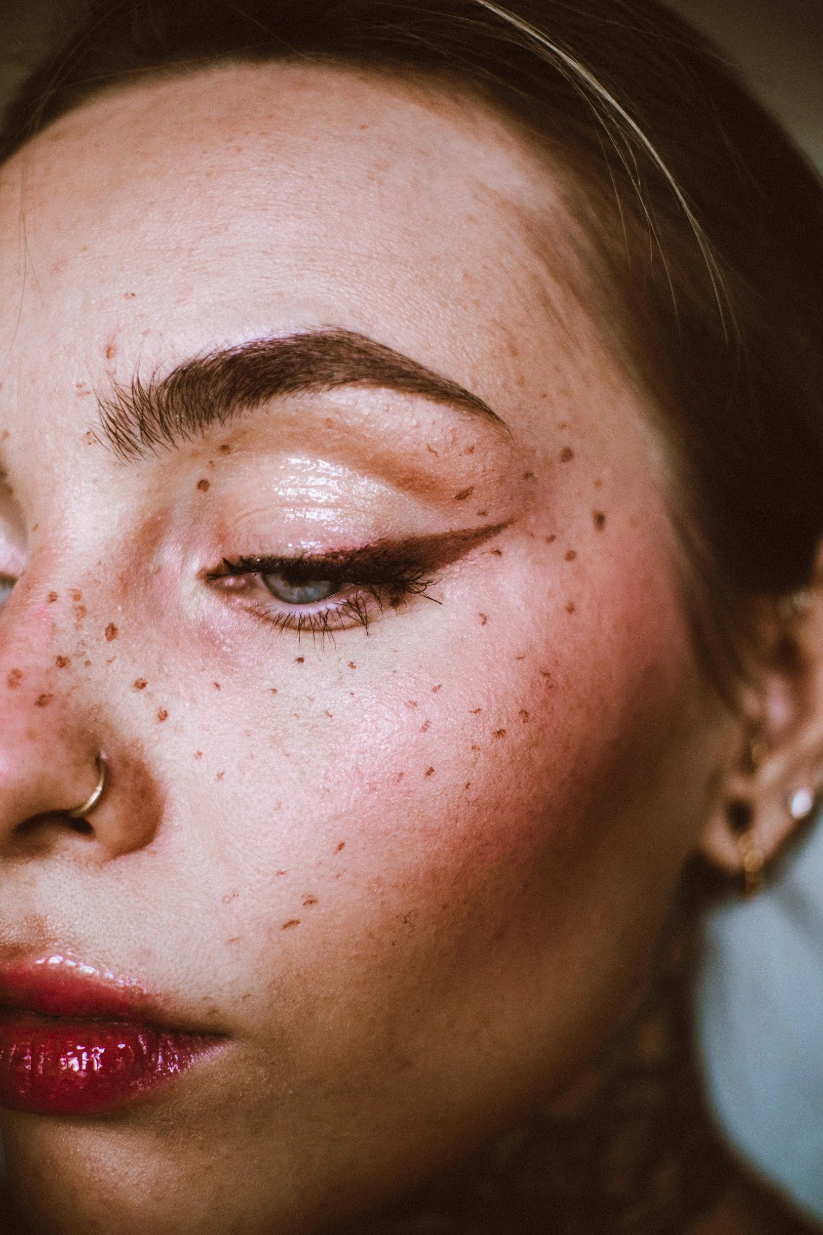 Close-up of a woman's face, showing her right eye closed, with dark brown eyebrows, PMU freckles, glossy red lips, and earrings.