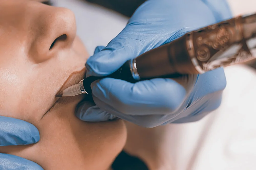Person receiving a cosmetic lip tattoo or permanent makeup procedure, with a technician in blue gloves applying ink to lips.