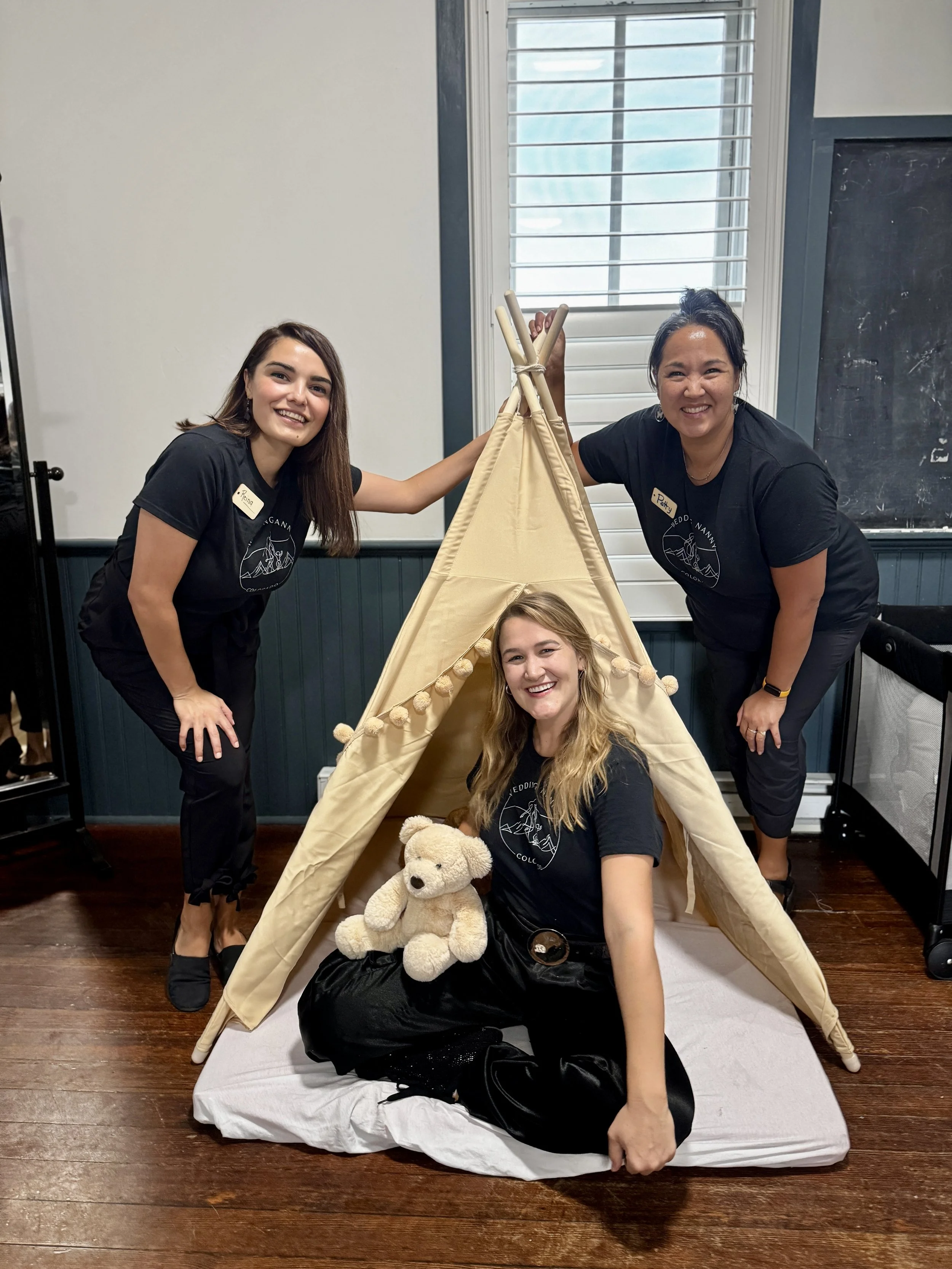 Four wedding nannies posing around a small beige teepee tent indoors, with one sitting on a white sheet with a teddy bear, two standing on the sides of the tent, and one resting her hand on the tent's peak. All are smiling and wearing black t-shirts.