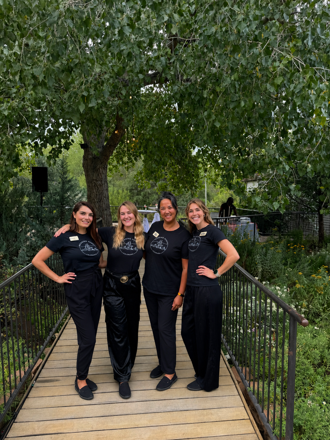 Four women dressed in black standing on a wooden pathway in a lush garden with a large tree overhead. They are smiling with their arms around each other.