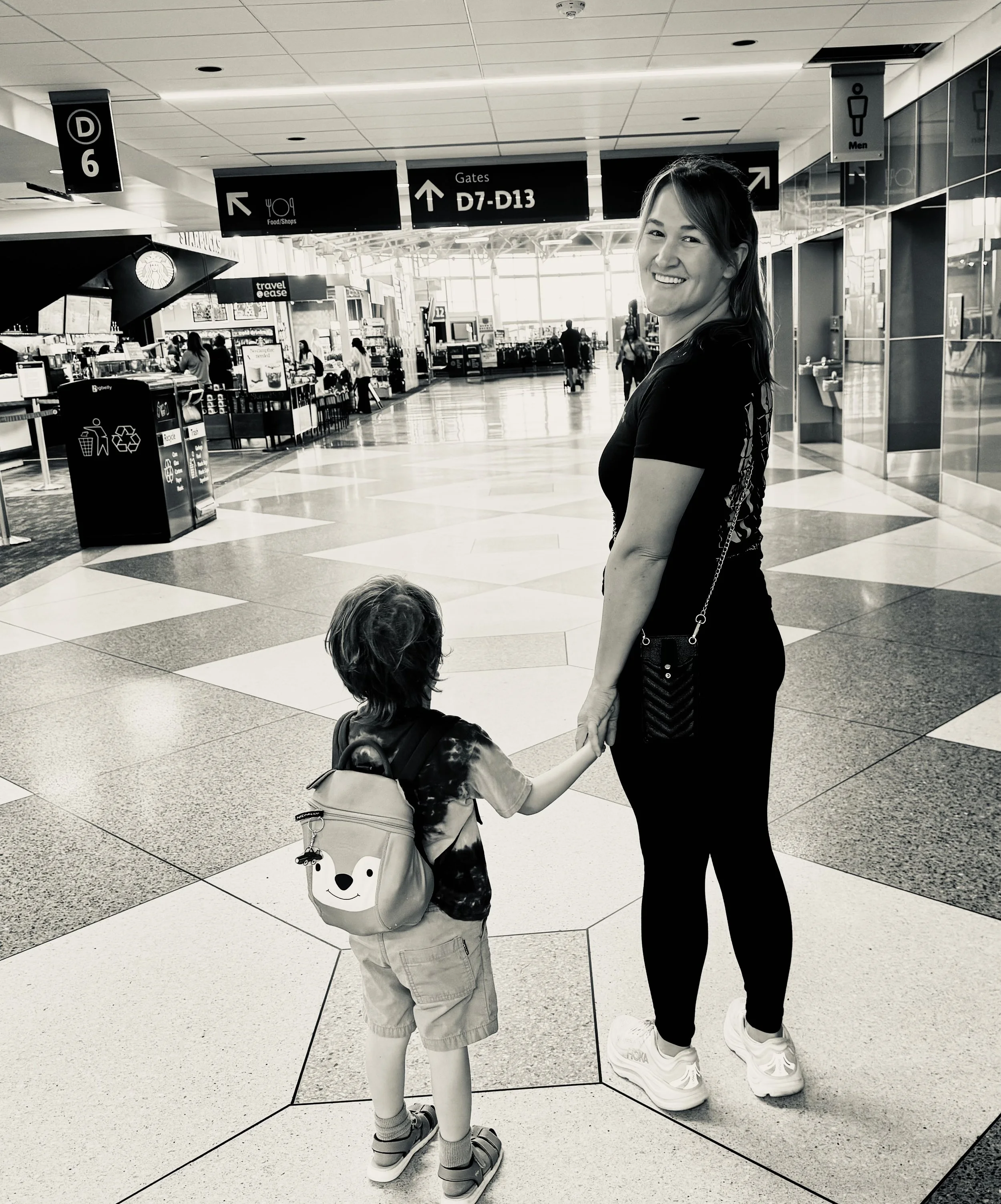 A woman and a young boy holding hands in an airport terminal, with signs overhead indicating gates D7-D13 and food shops.