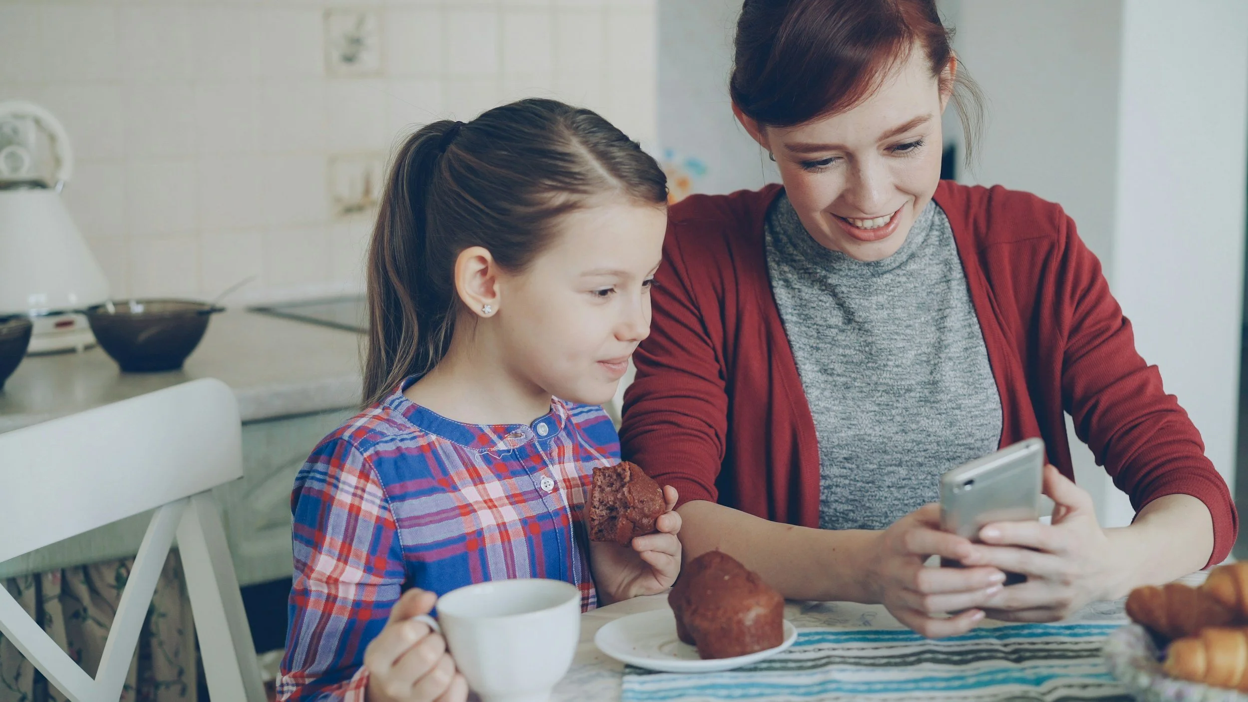 A woman and a young girl sitting together at a kitchen table, looking at a smartphone, with cups of coffee and baked goods on the table.