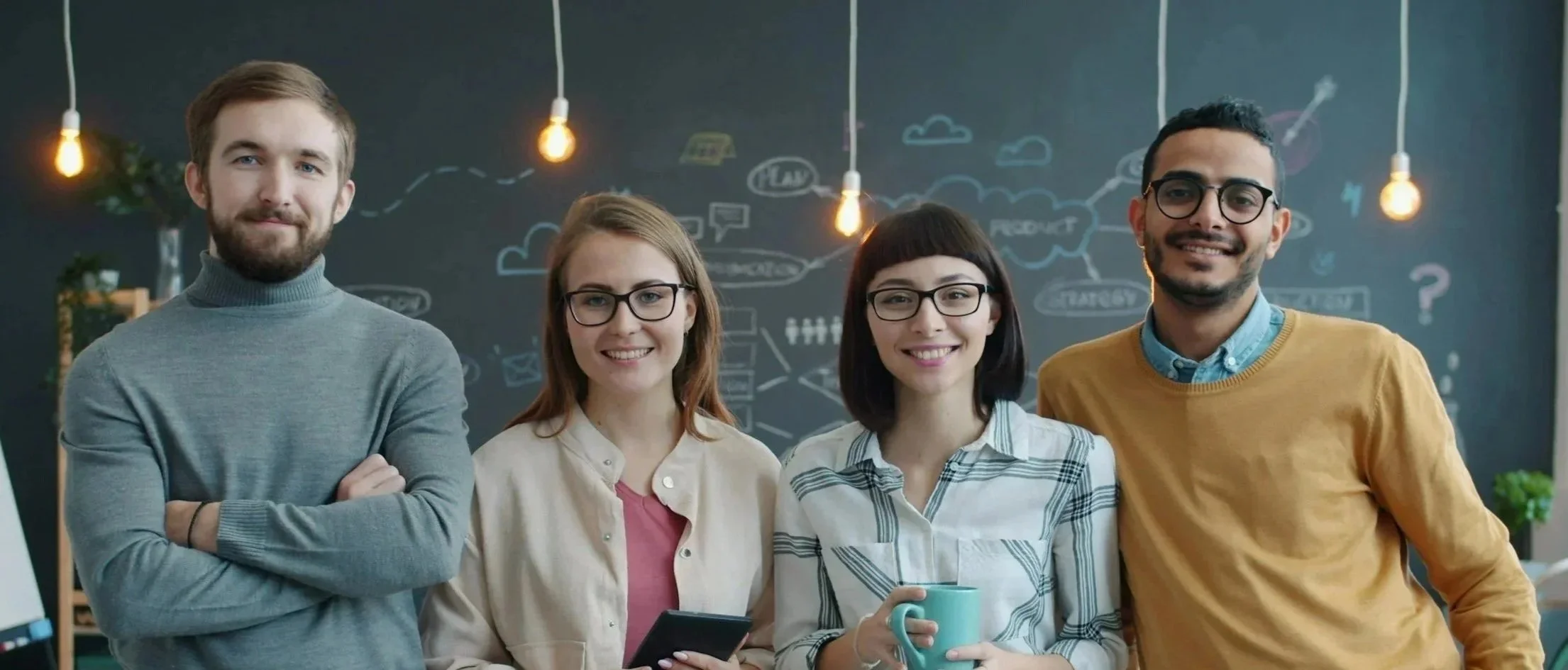 Four diverse young adults smiling together in a modern office, with a chalkboard wall filled with sketches and diagrams behind them.