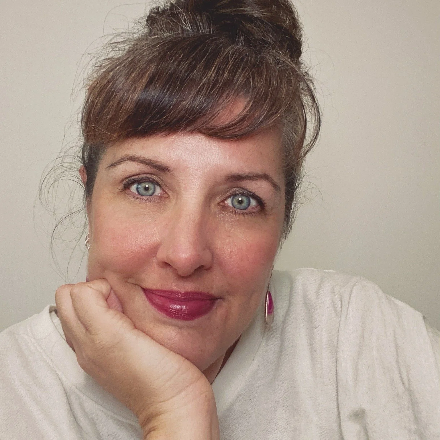 Close-up of a woman with short brown hair, blue eyes, and light skin, resting her chin on her hand and smiling softly, against a plain wall background.