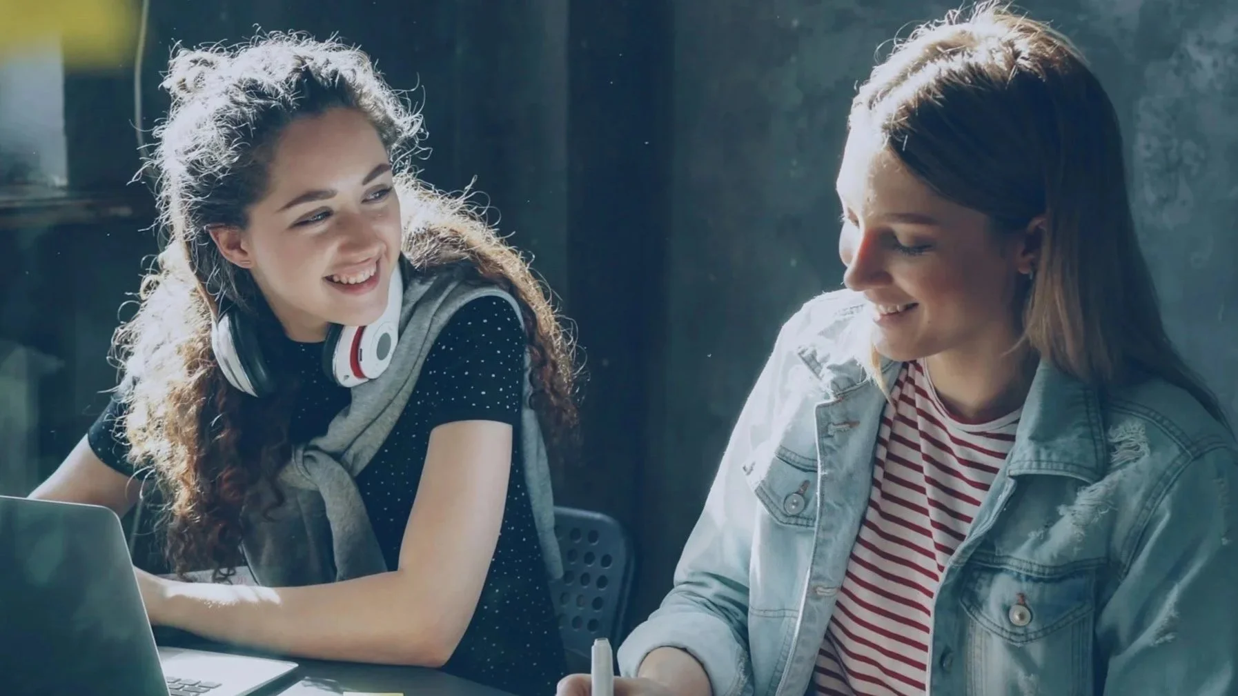 Two young women smiling and sitting together at a table, with one wearing headphones around her neck and the other holding a pen, engaged in conversation.