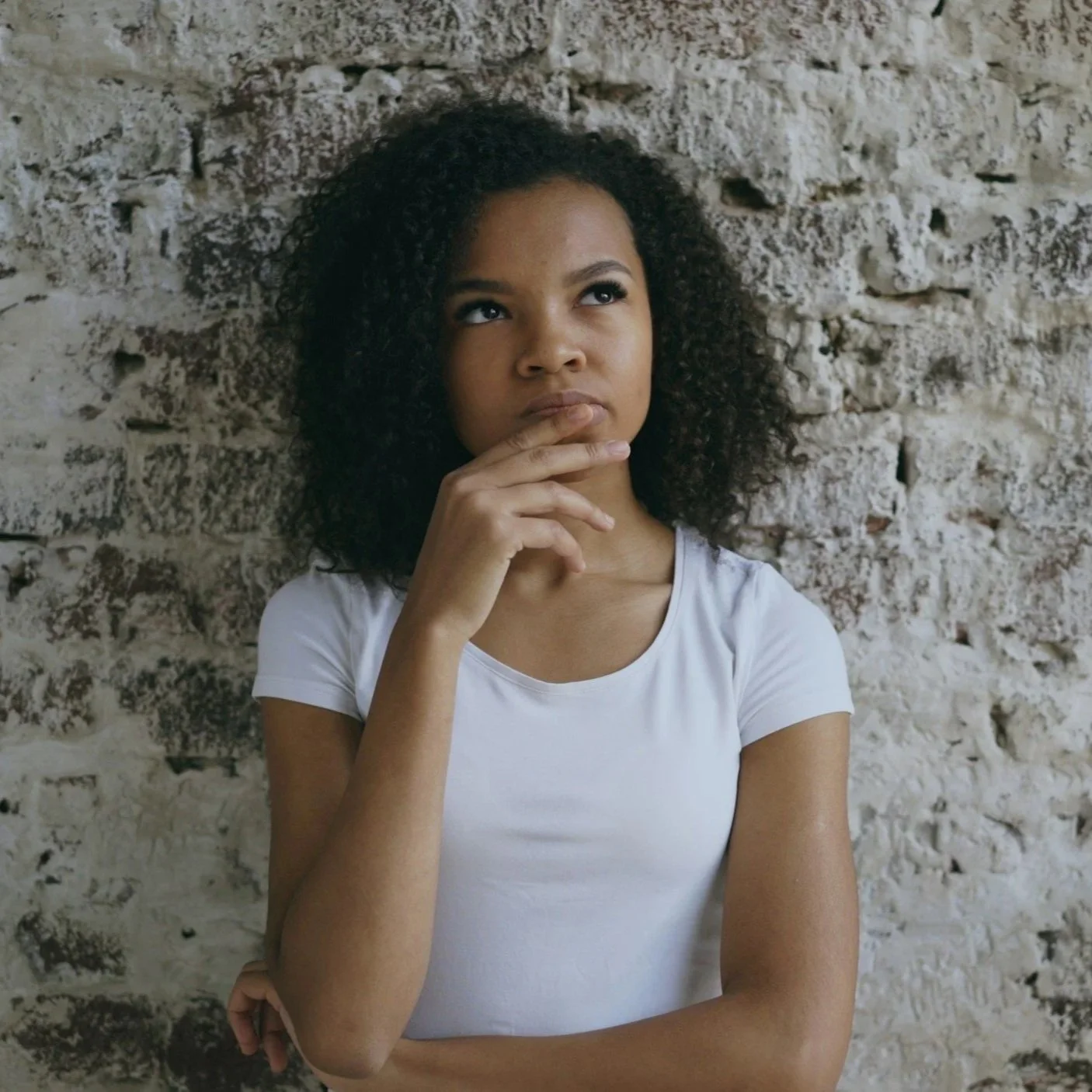 A young woman with curly hair wearing a white t-shirt, standing against a textured brick wall, with her hand resting near her chin and looking thoughtfully upward.