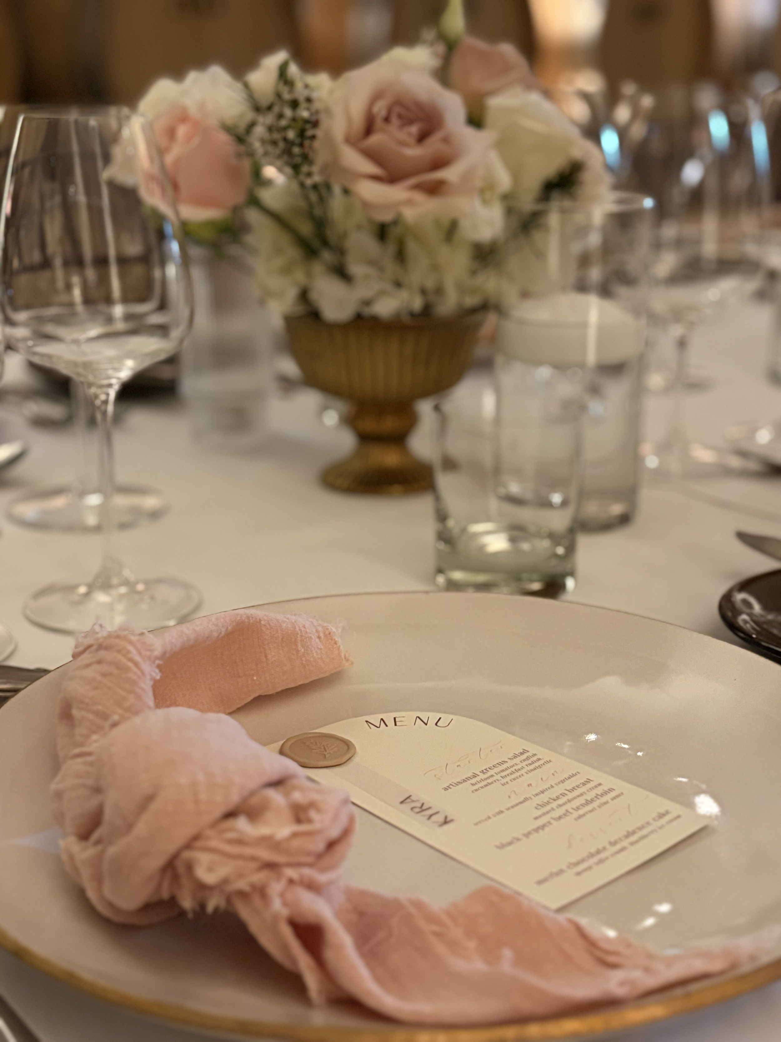 A table setting with a menu on a pink cloth napkin, a carved candle, and a floral centerpiece with roses and other flowers, surrounded by glassware and silverware.