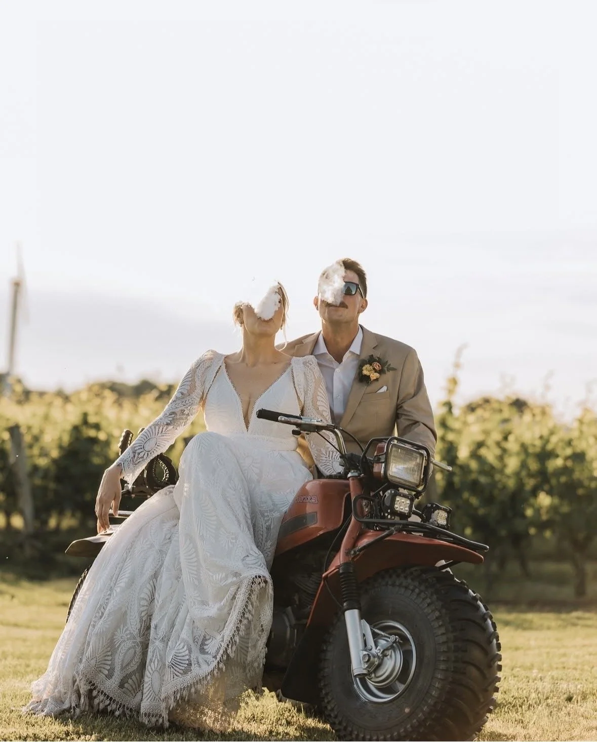 A bride and groom sitting on a three wheeler in a field, both blowing smoke, with a sunny sky and greenery in the background.