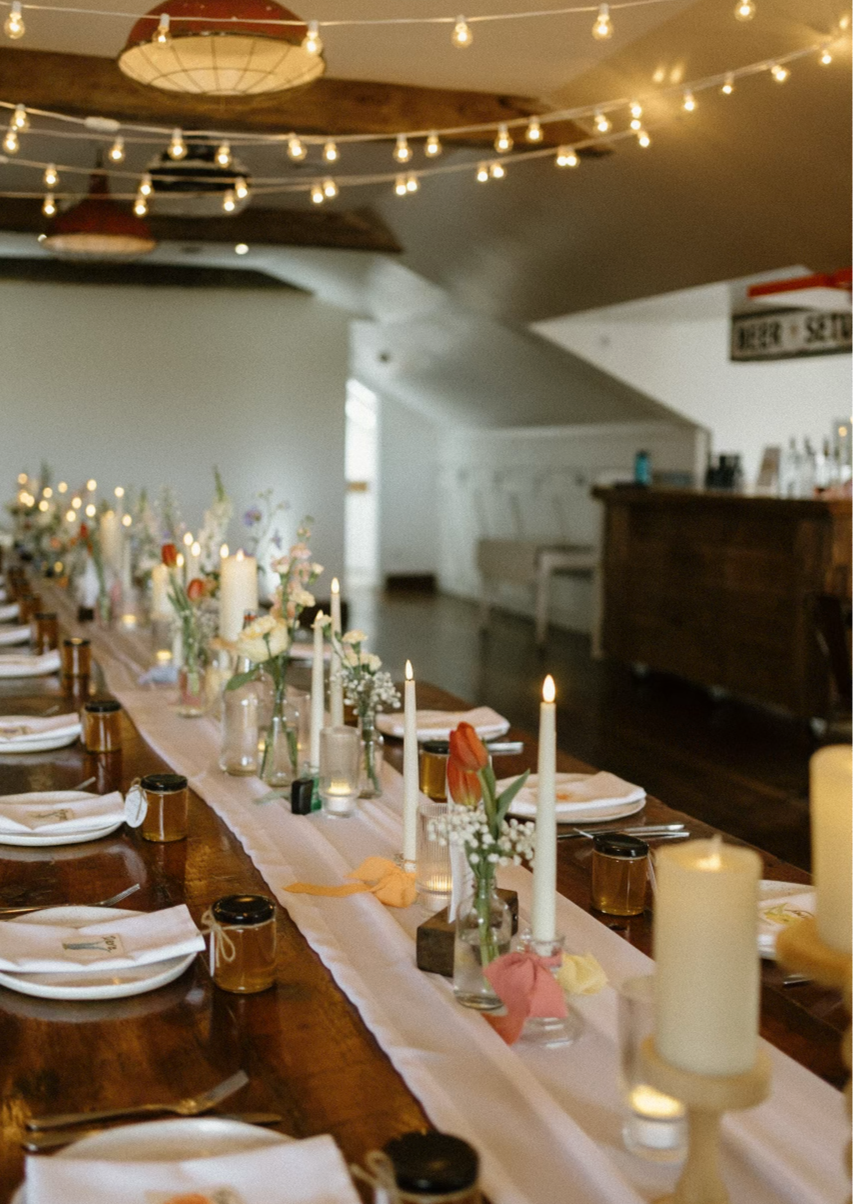 A long dining table decorated with flowers, candles, and place settings, illuminated by string lights overhead in a cozy indoor venue.
