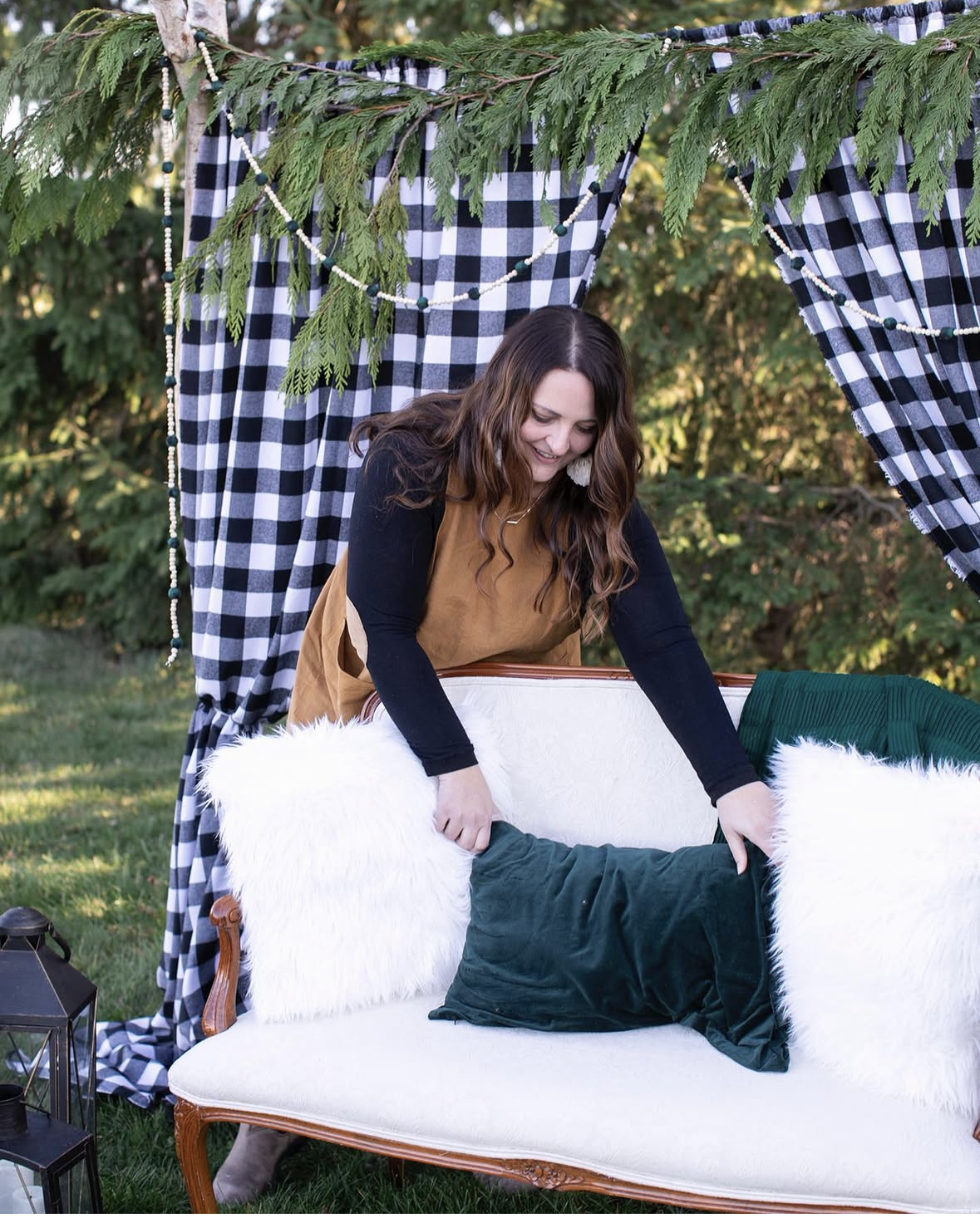 Beth arranging pillows on a white vintage sofa outdoors, with a backdrop of trees and greenery. The scene is decorated with a leafy canopy, checkered curtains, and hanging bead garland, creating a cozy outdoor setup.