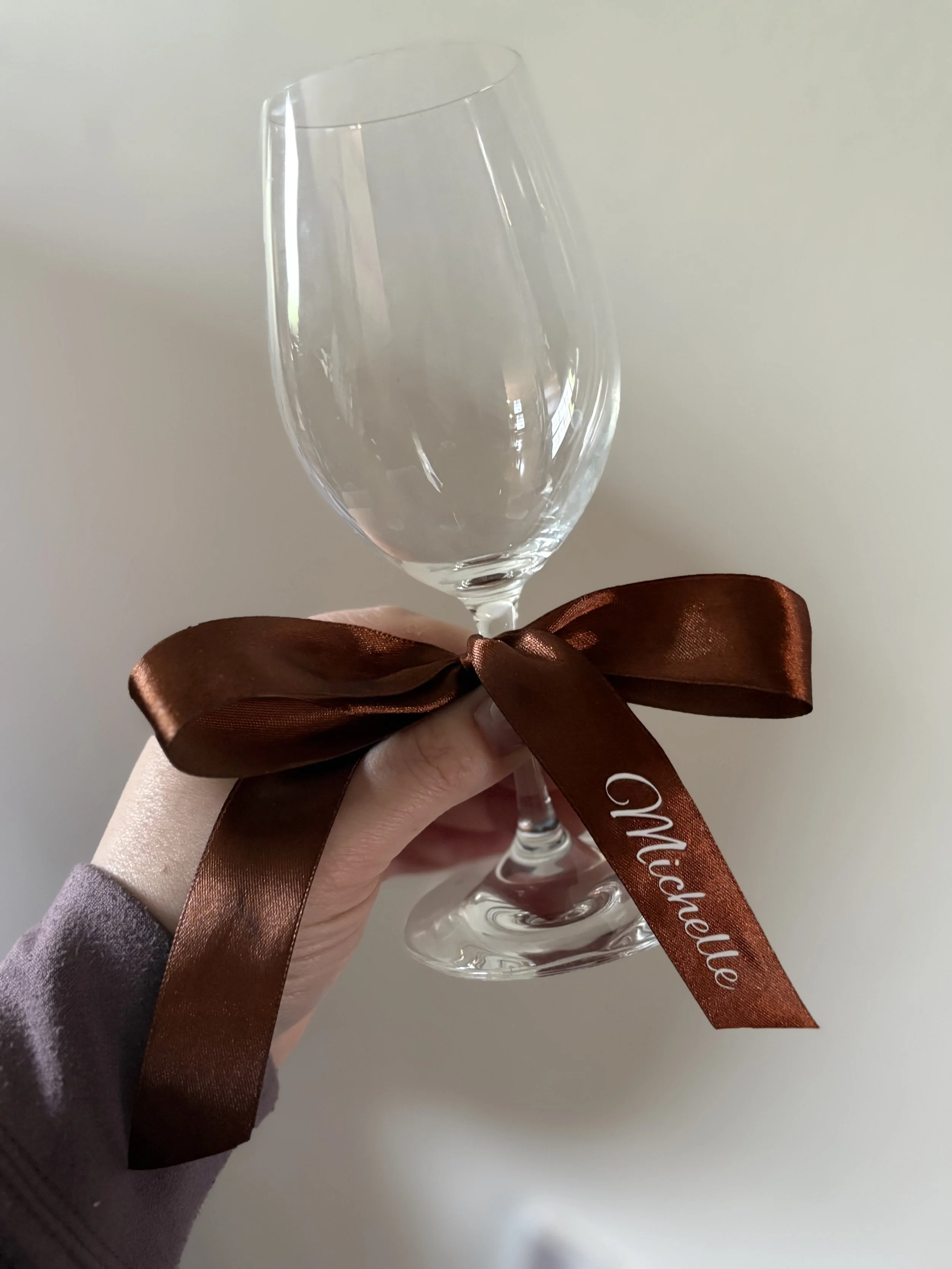A person's hand holding an empty wine glass decorated with a brown satin ribbon tied in a bow, with the word 'Michelle' written on the ribbon.