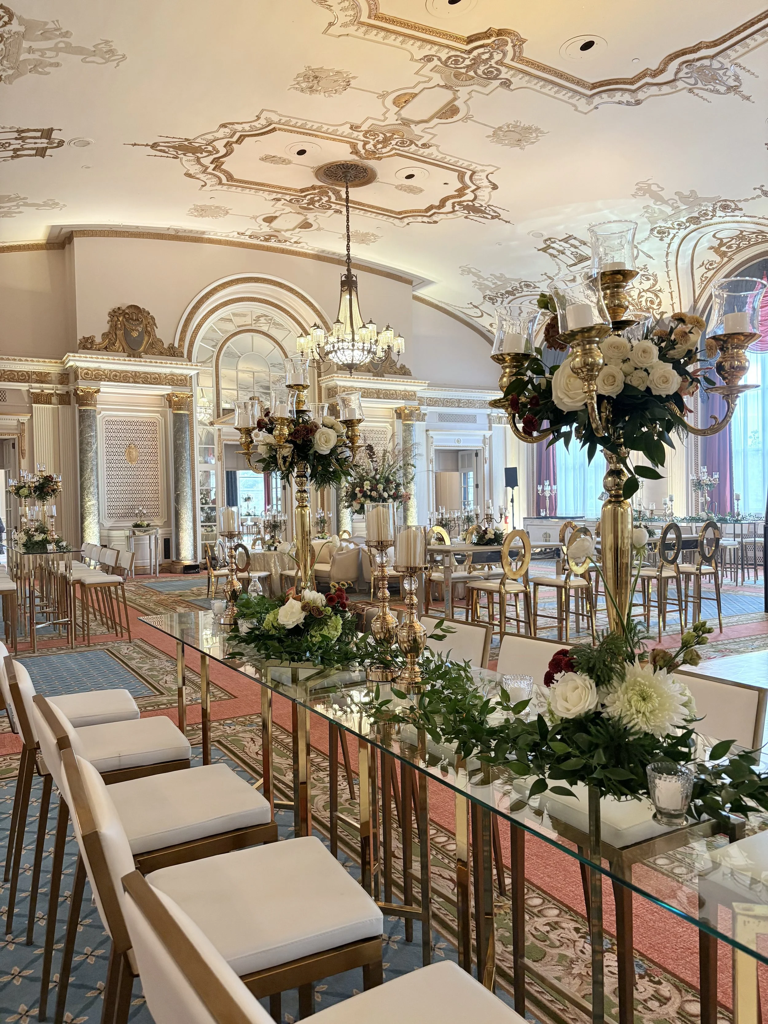 Elegant banquet or wedding reception hall with white and gold decor, large floral arrangements, gold candle holders with candles, and chandeliers hanging from ornately decorated ceilings.