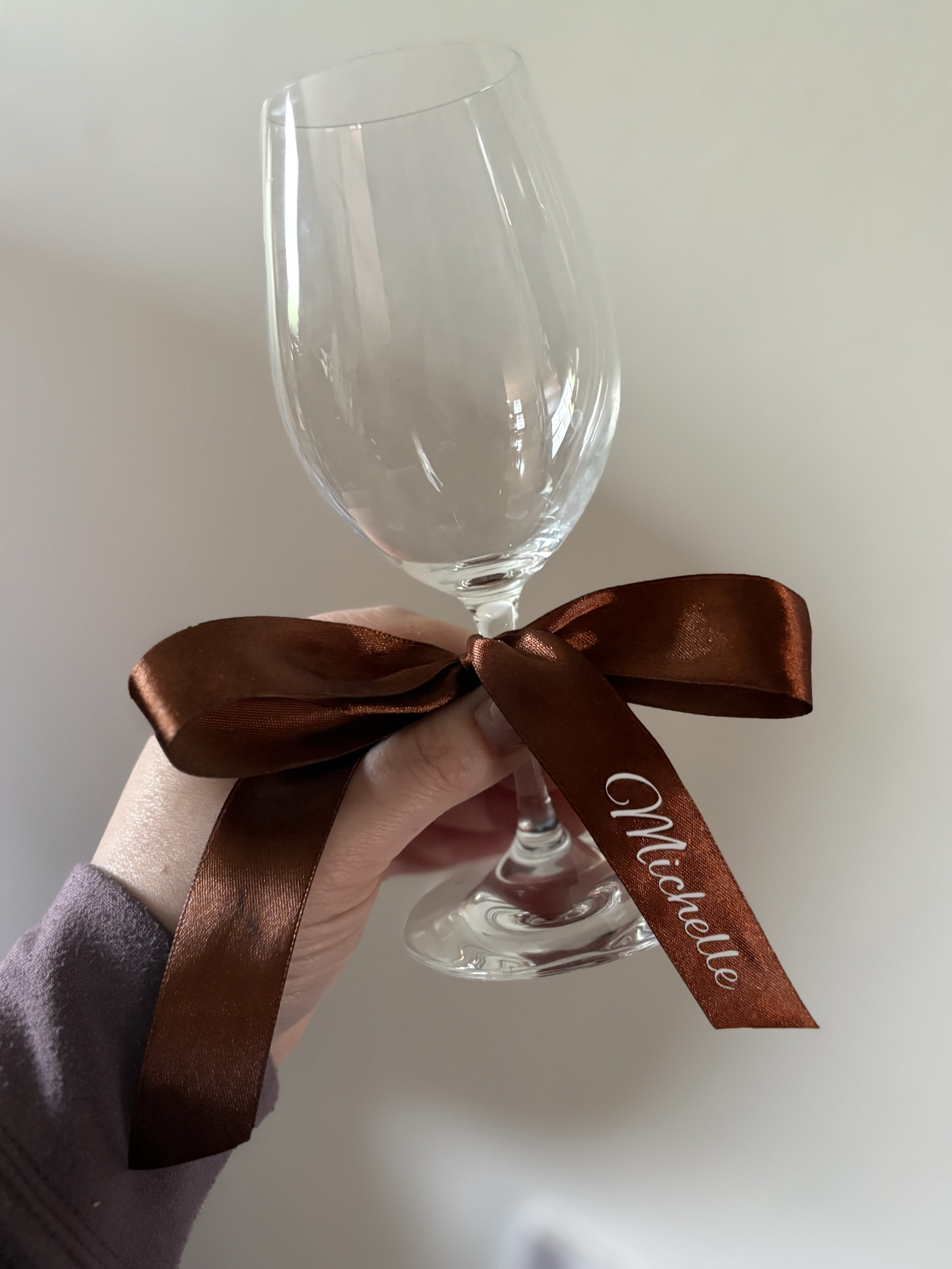 A hand holding an empty wine glass with a brown satin ribbon tied around its stem, with ''Michelle'' written on the ribbon, against a plain background.