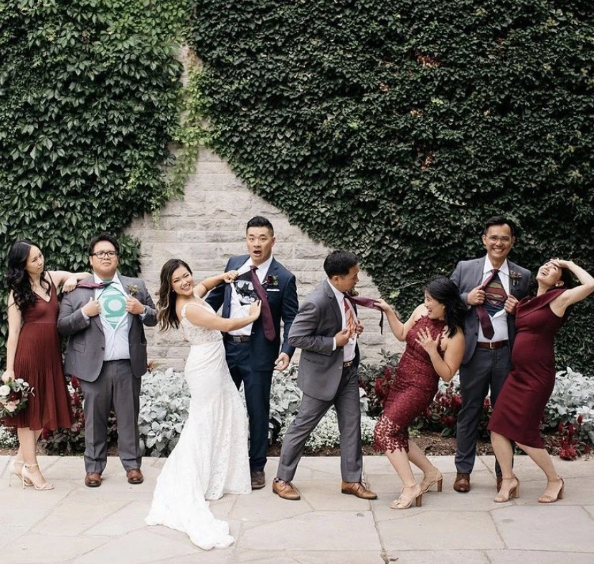 Group of eight diverse wedding guests posing playfully in front of a green leafy wall, with some pulling on the ties of others, all smiling and dressed in formal attire.