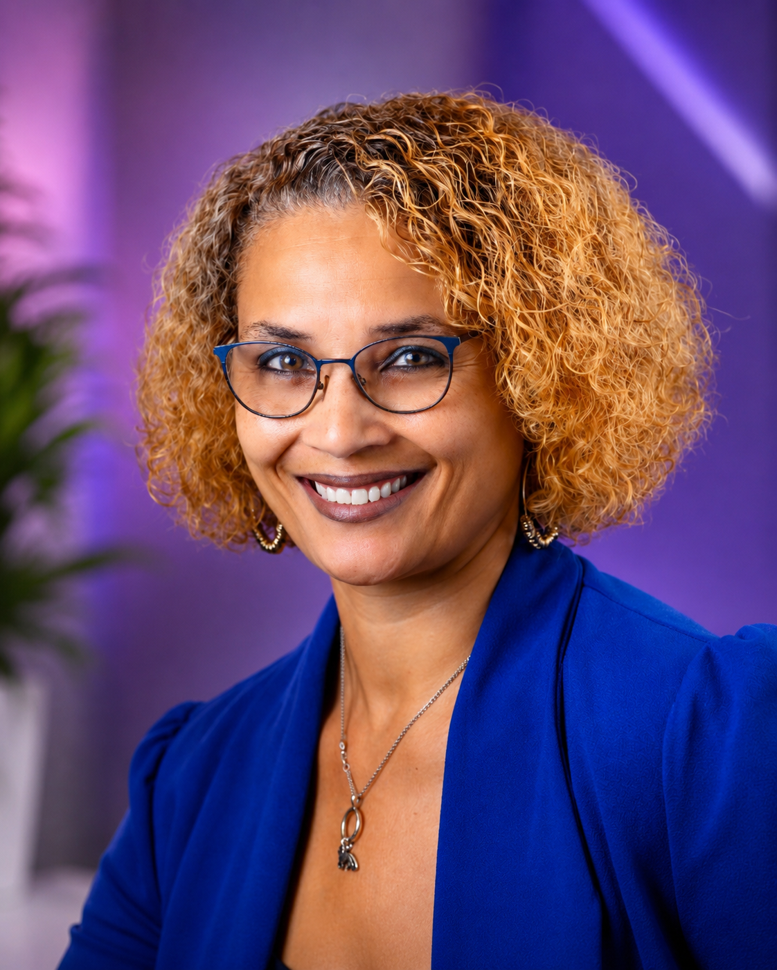A woman with curly blonde hair, glasses, and brown skin smiling. She is wearing a blue blazer, earrings, and a necklace.