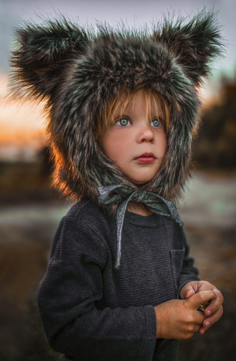 A young child with blue eyes and light brown hair, wearing a dark sweater and a large furry hat with ear flaps, standing outdoors during sunset.
