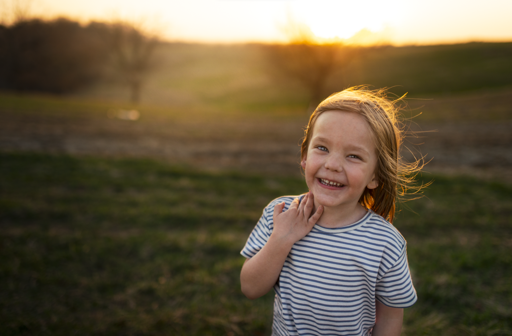 A young girl with red hair smiling outdoors during sunset, wearing a striped t-shirt.