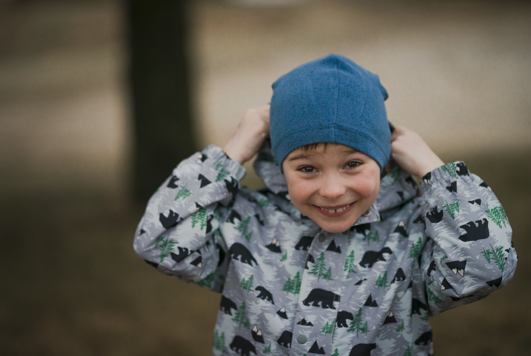 A young boy wearing a blue beanie and a patterned raincoat with bears and trees, smiling and adjusting his hat with both hands outdoors.