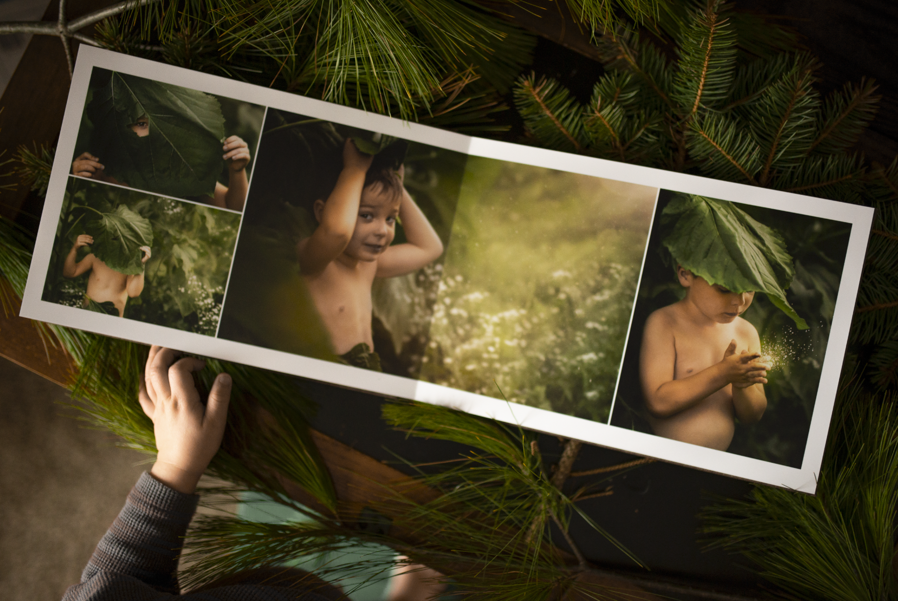 A photo collage of a young boy, shirtless, in nature, holding large leaves to his face and wearing a leaf on his head, set on a rustic wooden table surrounded by pine branches.