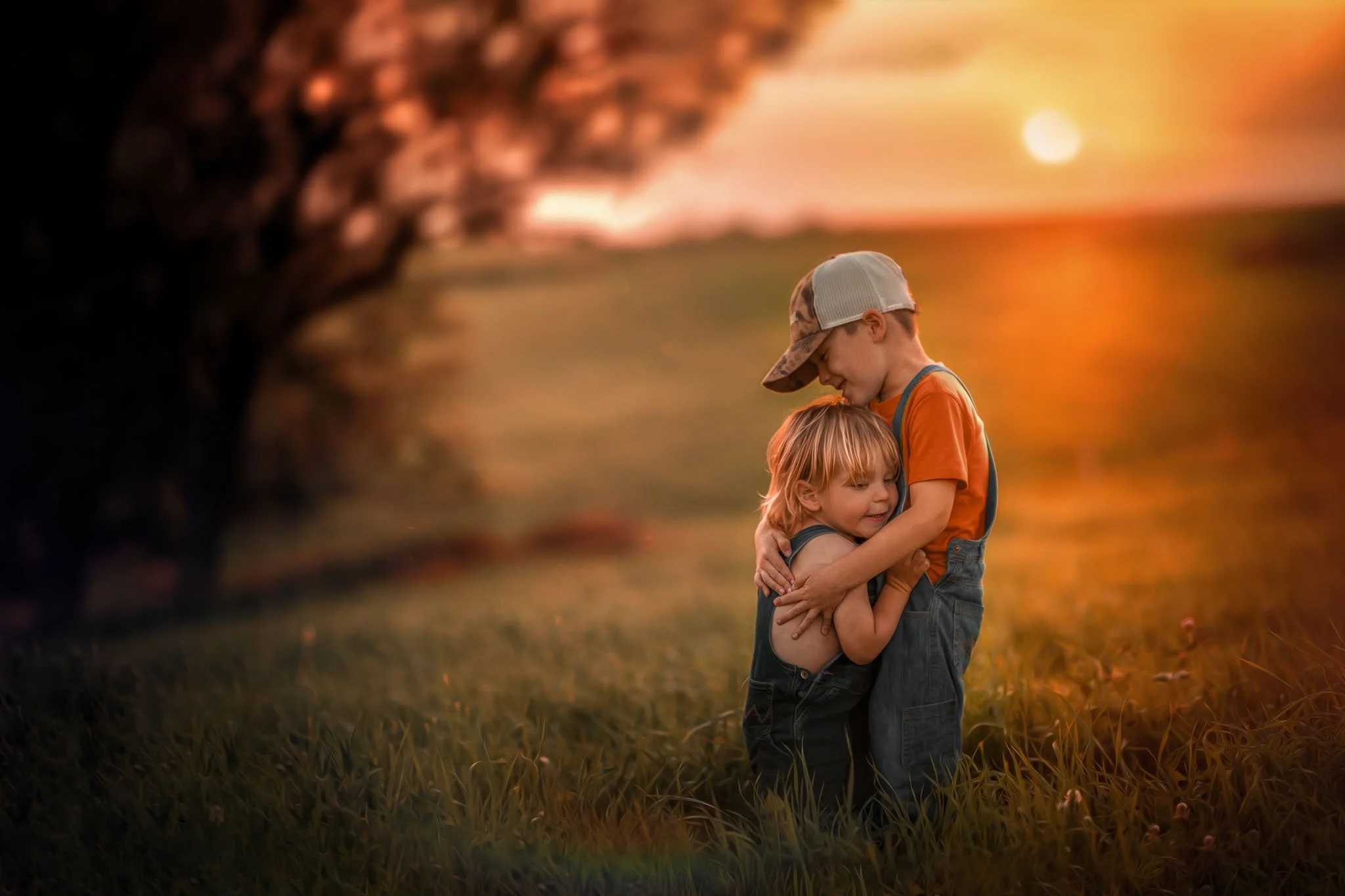 Two children, a boy and a girl, embracing each other outdoors during sunset. The boy is wearing an orange shirt, a cap, and suspenders. The girl has red hair and is smiling. The background is a warm, glowing sunset with a blurred landscape.