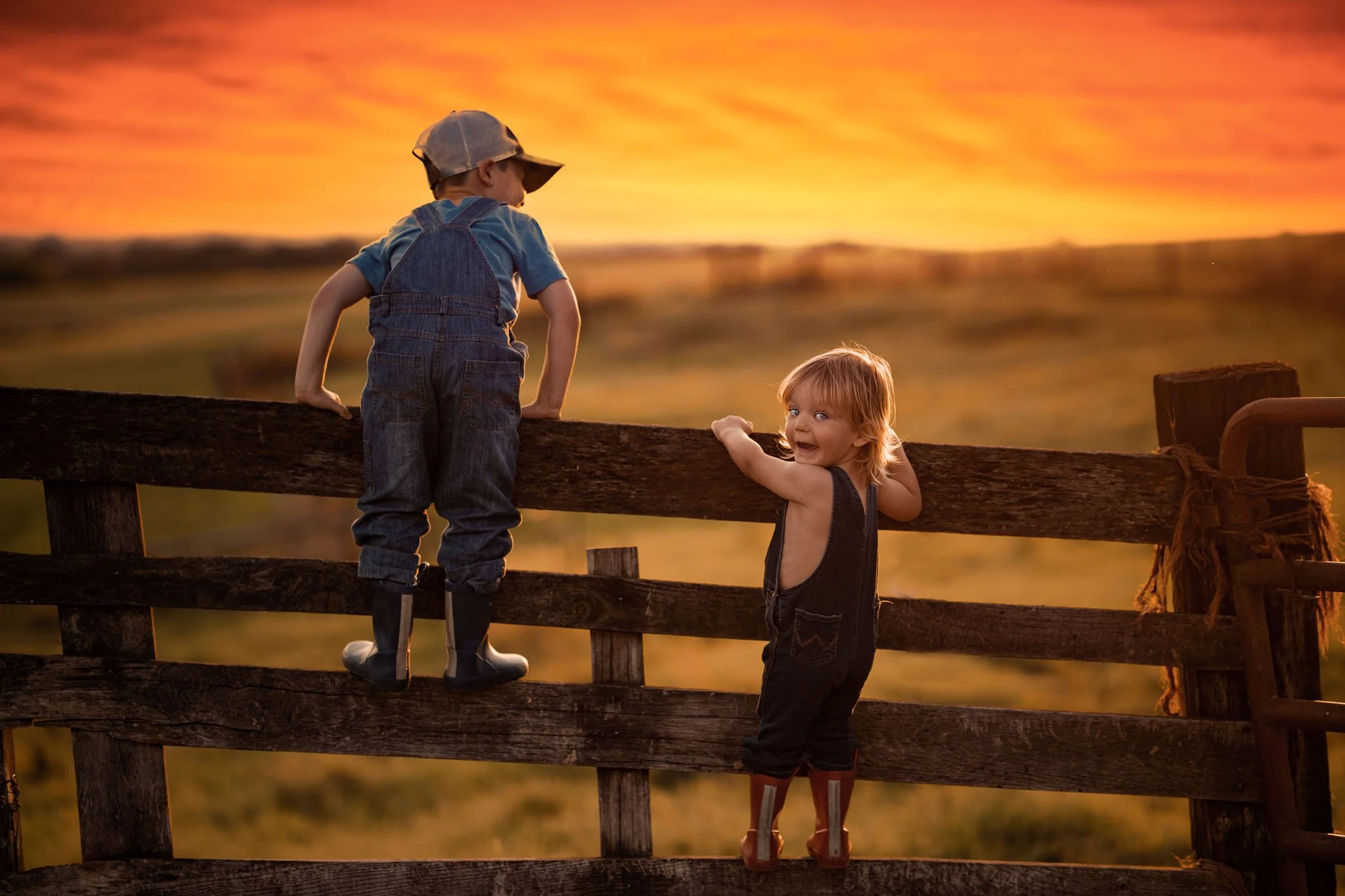 Two young children, a boy and a girl, standing on a wooden fence and looking at a vibrant sunset sky with shades of orange and pink.