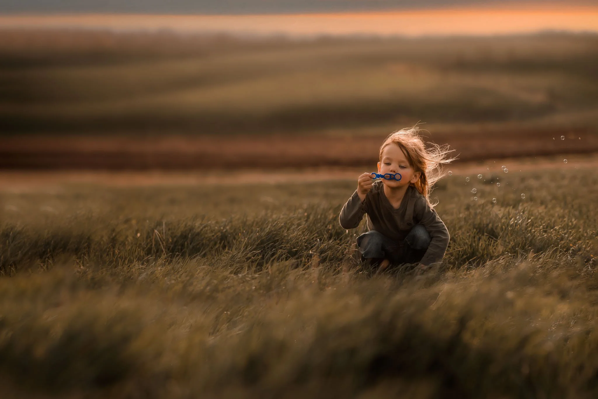 A young girl with red hair playing with bubbles in a field during sunset.