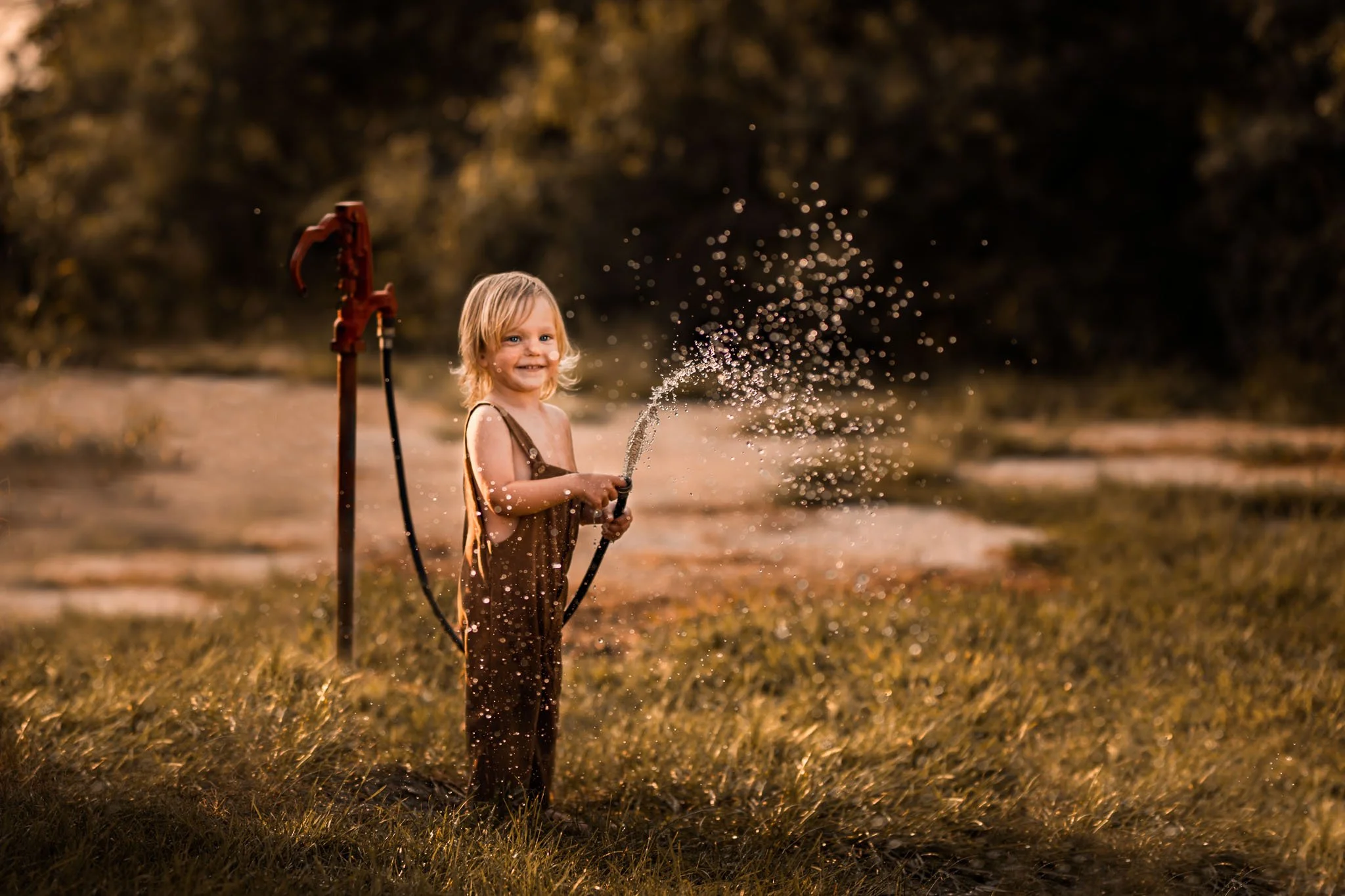 A young girl with blonde hair smiling and playing with a garden hose, spraying water in a backyard during golden hour. She is wearing a brown outfit and standing on grass with trees in the background.