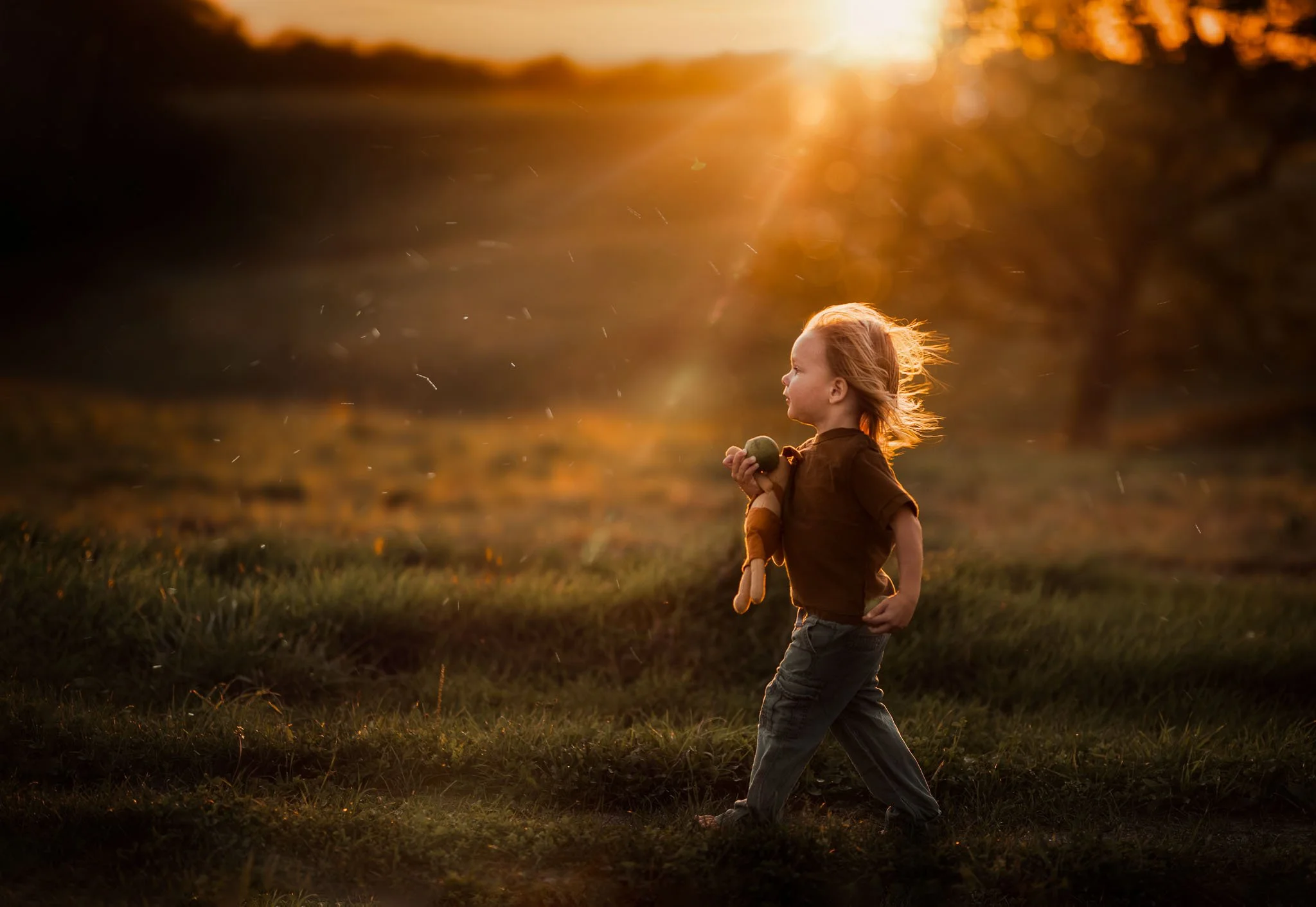 A young child with red hair walking outdoors at sunset, holding a small ball in one hand, with tall grass and trees in the background.