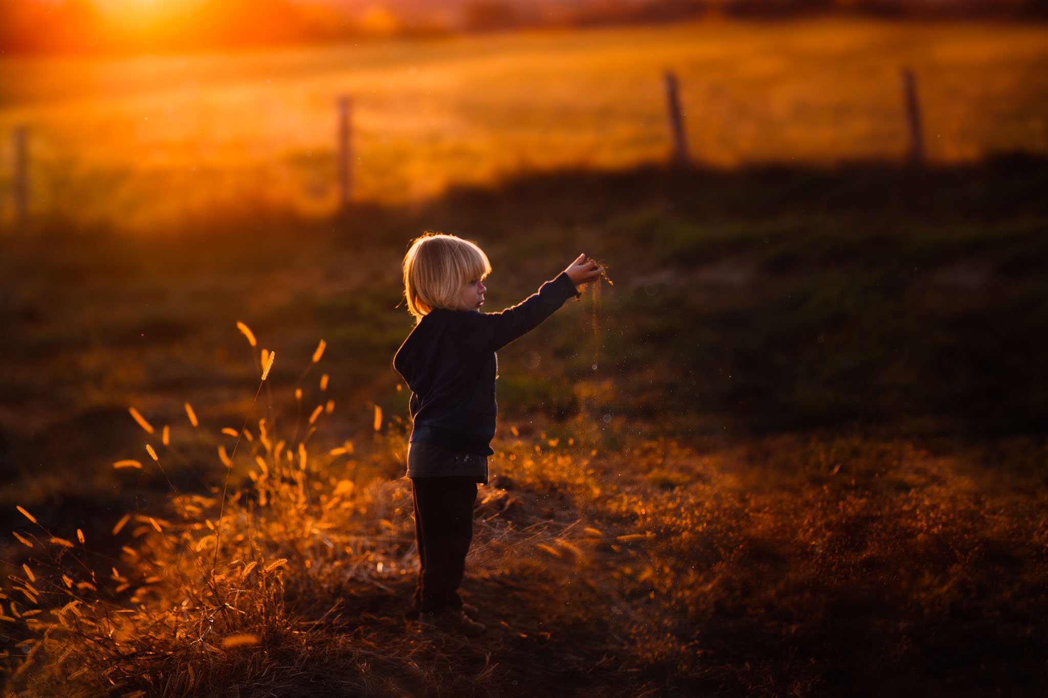 A young child with blonde hair playing with fire sparks outdoors during sunset, with a fence in the background.