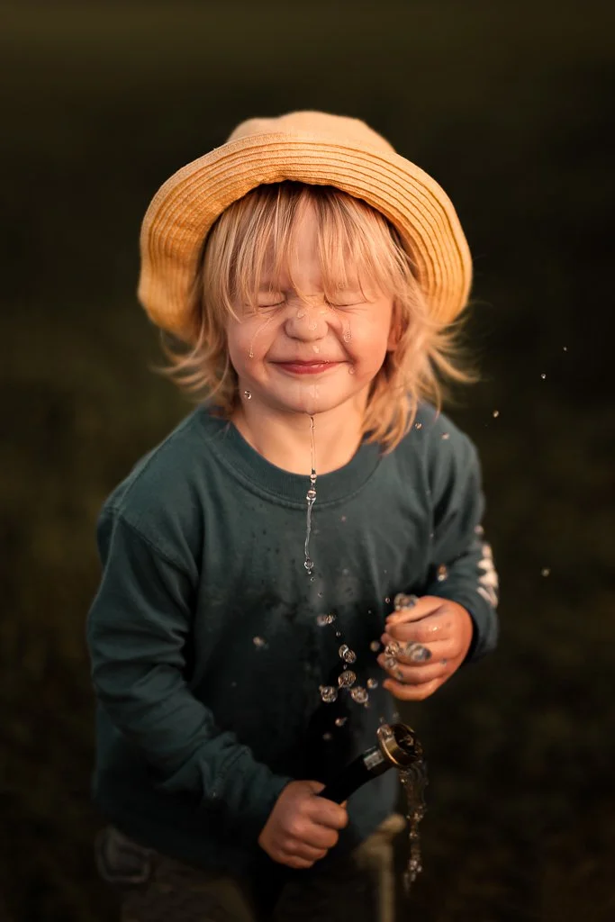 A young child with blond hair, wearing a straw hat and a dark green long-sleeve shirt, is happily playing with a hose outdoors in a grassy area during the daytime.