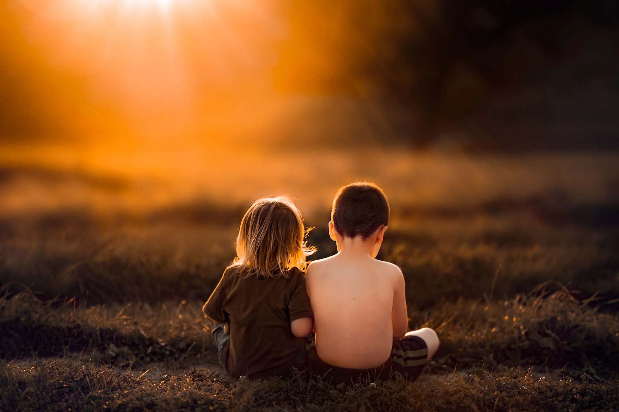 A boy and a girl sit together on the grass at sunset, watching the colorful sky.