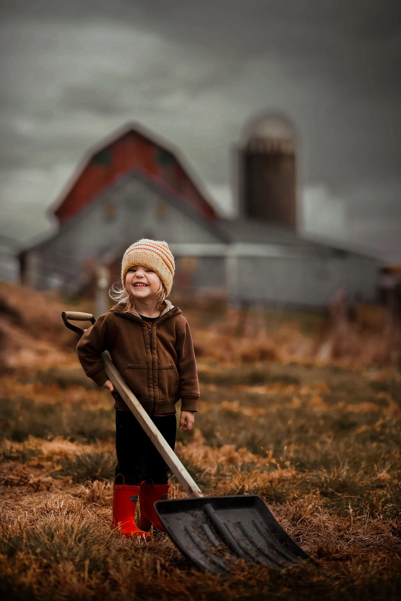 A young girl standing in a field of dry grass, wearing a cozy brown jacket, a knitted hat, red rain boots, and holding a shovel. She is smiling. In the background, there is a barn and a silo under a cloudy, overcast sky.