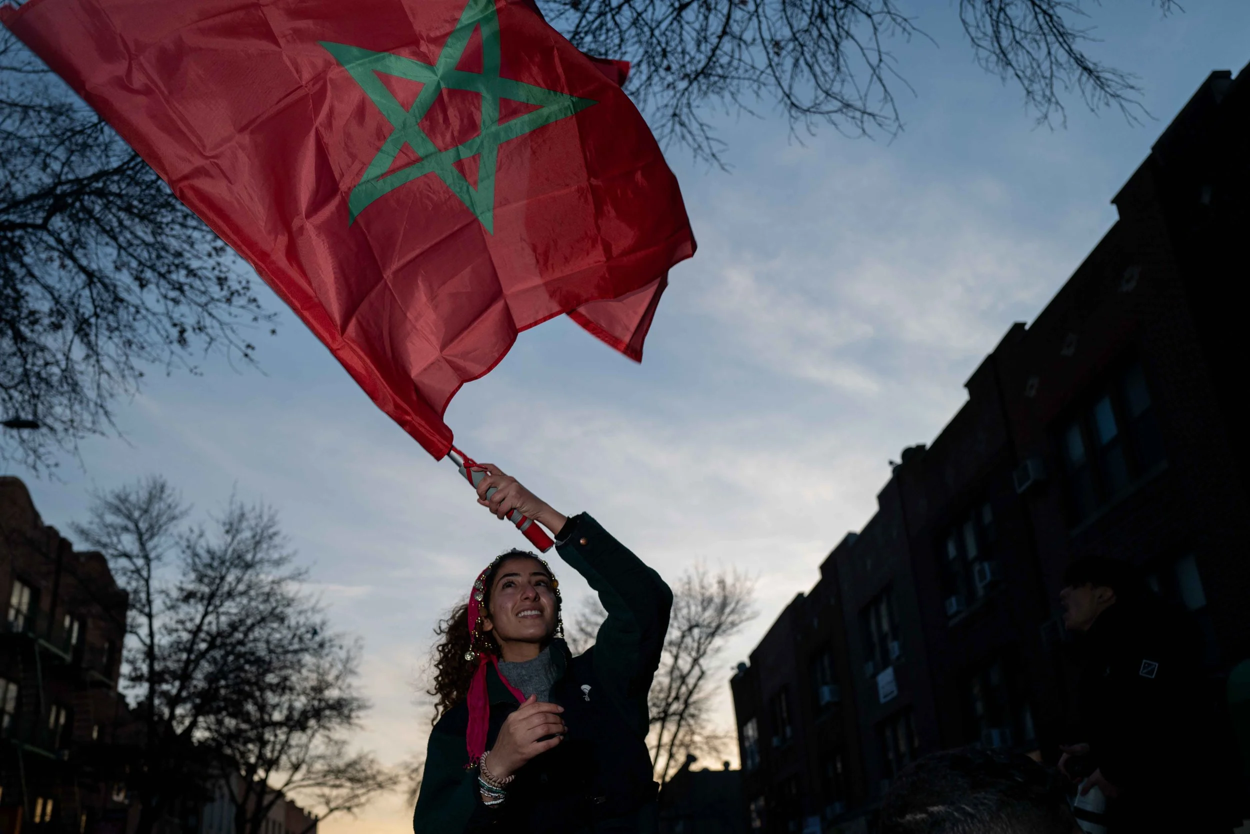 A woman is holding up a Moroccan flag outdoors during dusk, with bare trees and buildings visible in the background.